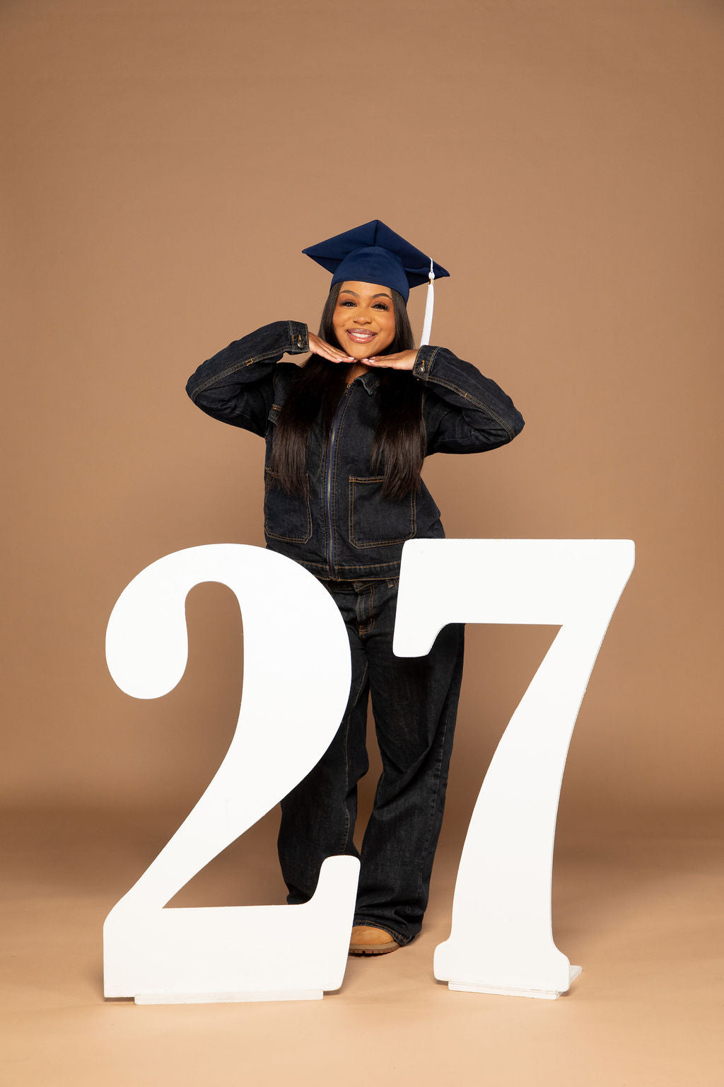 A young woman wearing a dark blue graduation cap and a dark denim jacket and jeans stands behind two large white numbers '27' on a solid tan studio background.