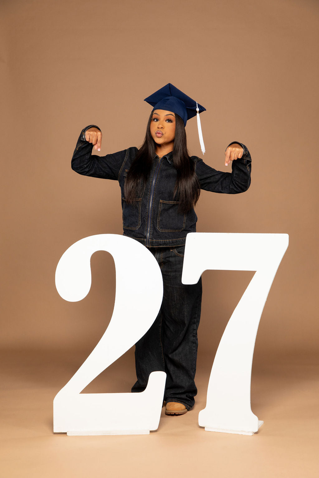 A young woman wearing a dark denim jacket, jeans, and a navy blue graduation cap stands behind two large white numbers '27' on a plain tan studio background.
