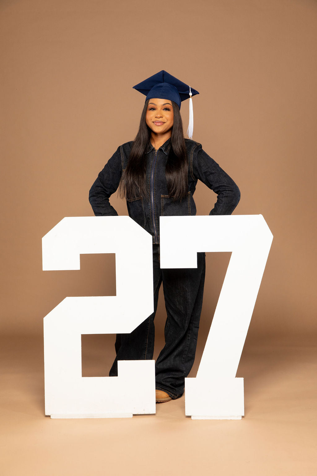 A young woman in a dark navy blue graduation gown and cap stands behind two large white numbers '27' on a solid tan studio background.