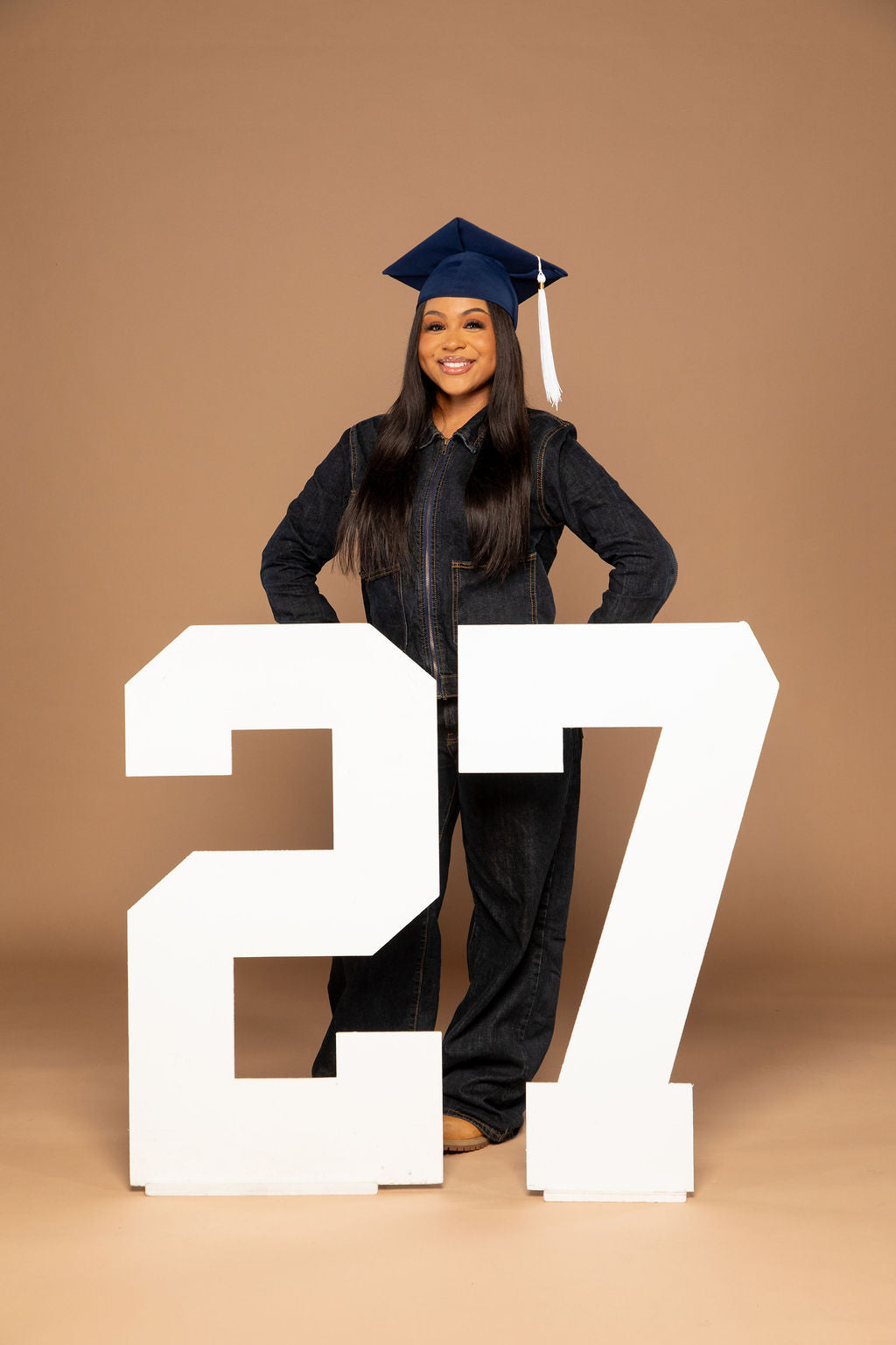 A young woman in a black graduation gown and cap stands behind large white numbers '27' against a solid tan studio background.