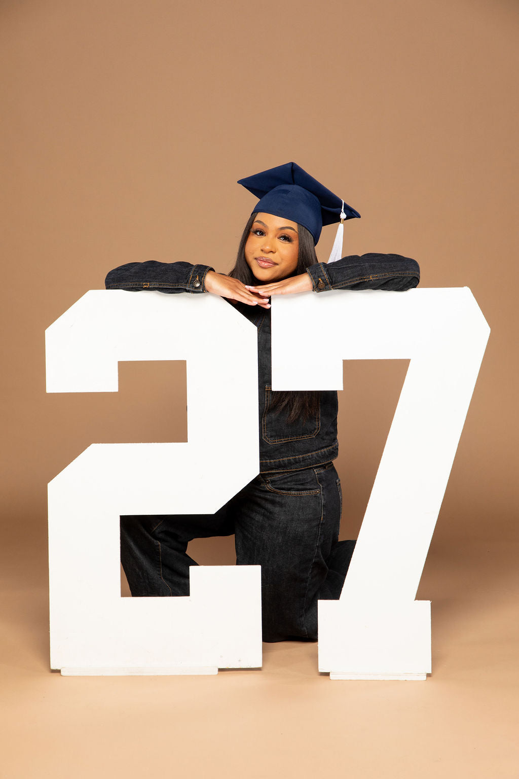 A graduate wearing a navy blue graduation cap and dark denim clothing kneels behind a large white number 27 prop against a solid tan studio background. Available at collageandwood.com.