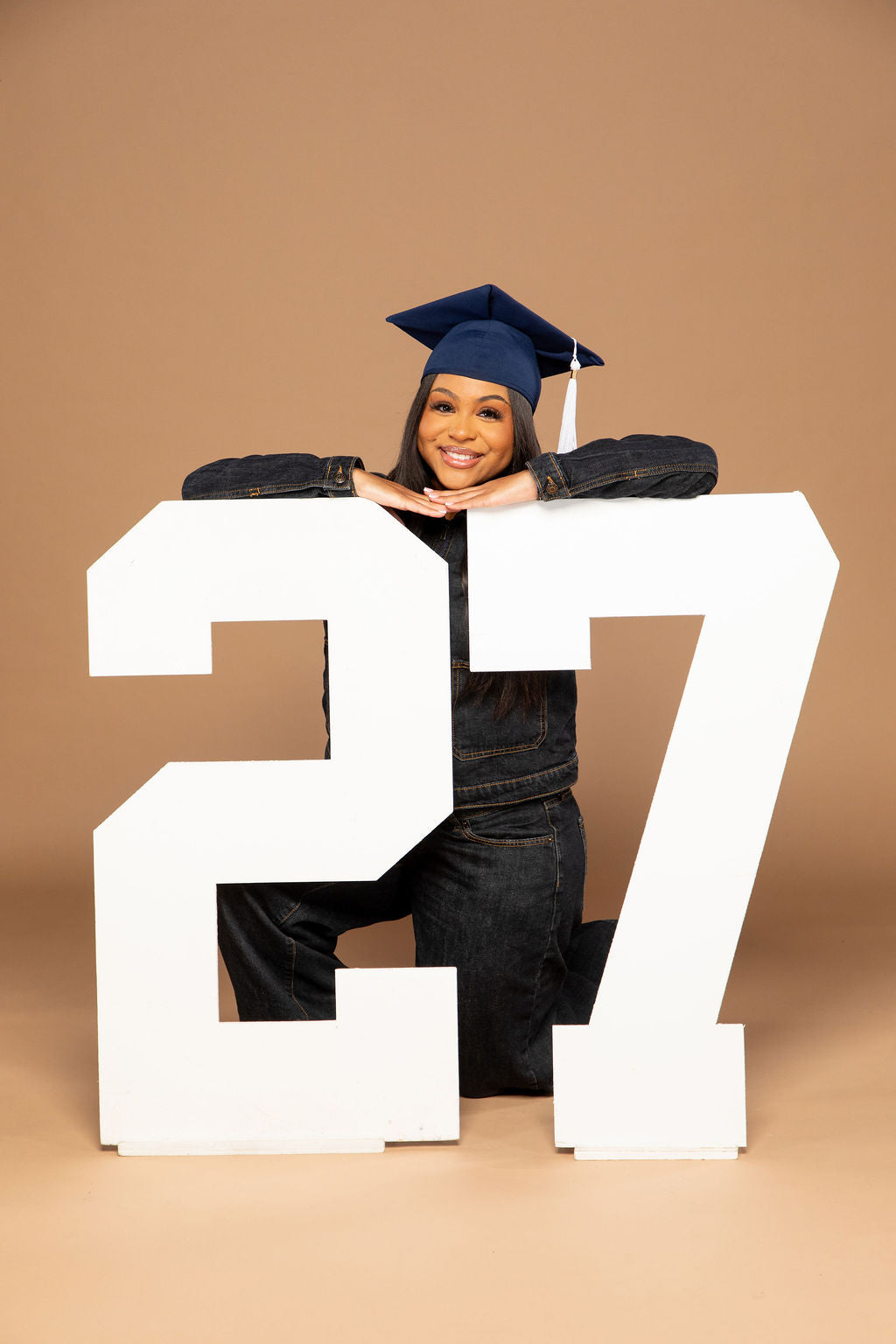 A smiling young woman in a navy blue graduation cap and dark denim outfit kneels behind oversized white numbers '27' on a warm tan studio background.