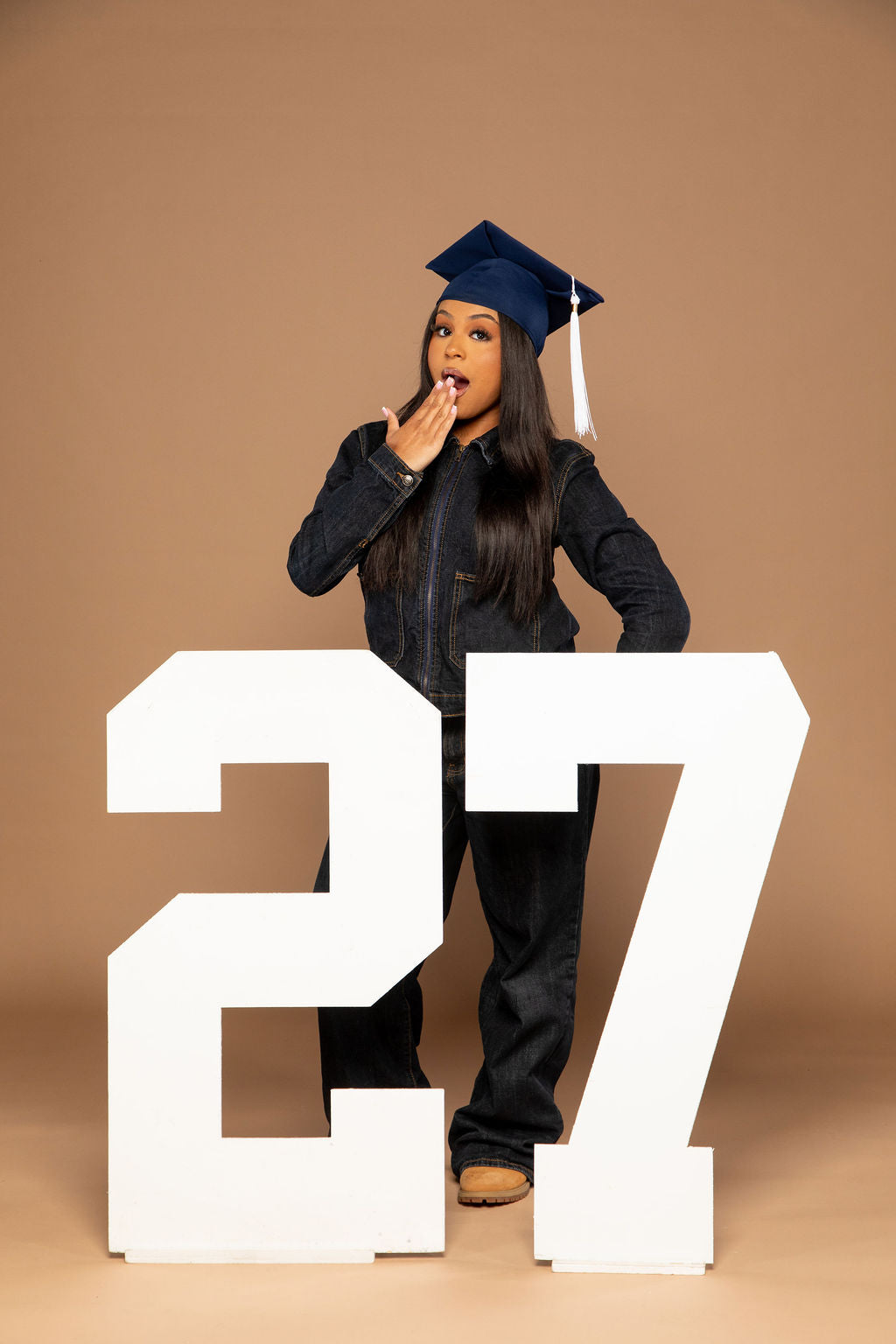 A woman in a dark denim graduation outfit and cap stands behind oversized white numbers '27' against a solid tan studio background.