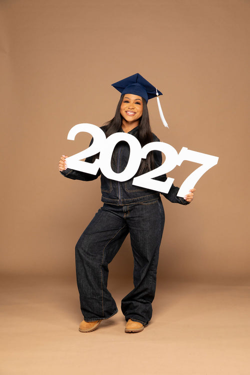 A smiling young woman in a navy blue denim outfit, dark jeans, tan shoes, and a navy blue graduation cap holds a large white '2027' sign against a solid tan studio backdrop. Available at collageandwood.com.