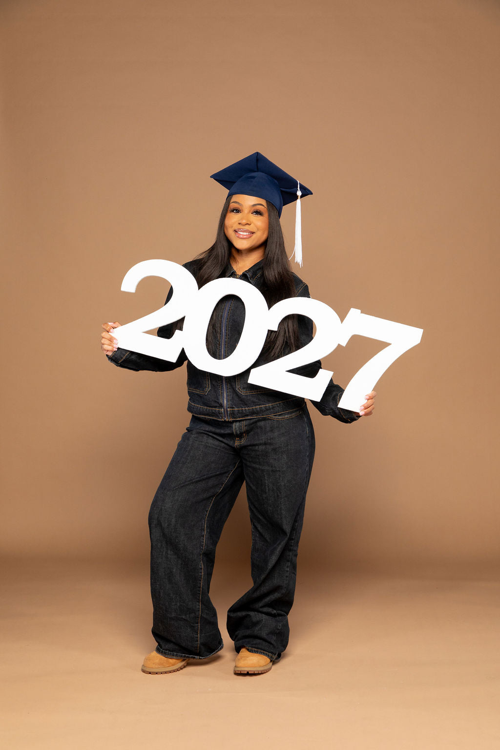 A woman wearing a navy blue graduation cap and dark denim jeans stands against a solid tan studio background, holding a large white 3D cutout of the numbers '2027'. Available at collageandwood.com.