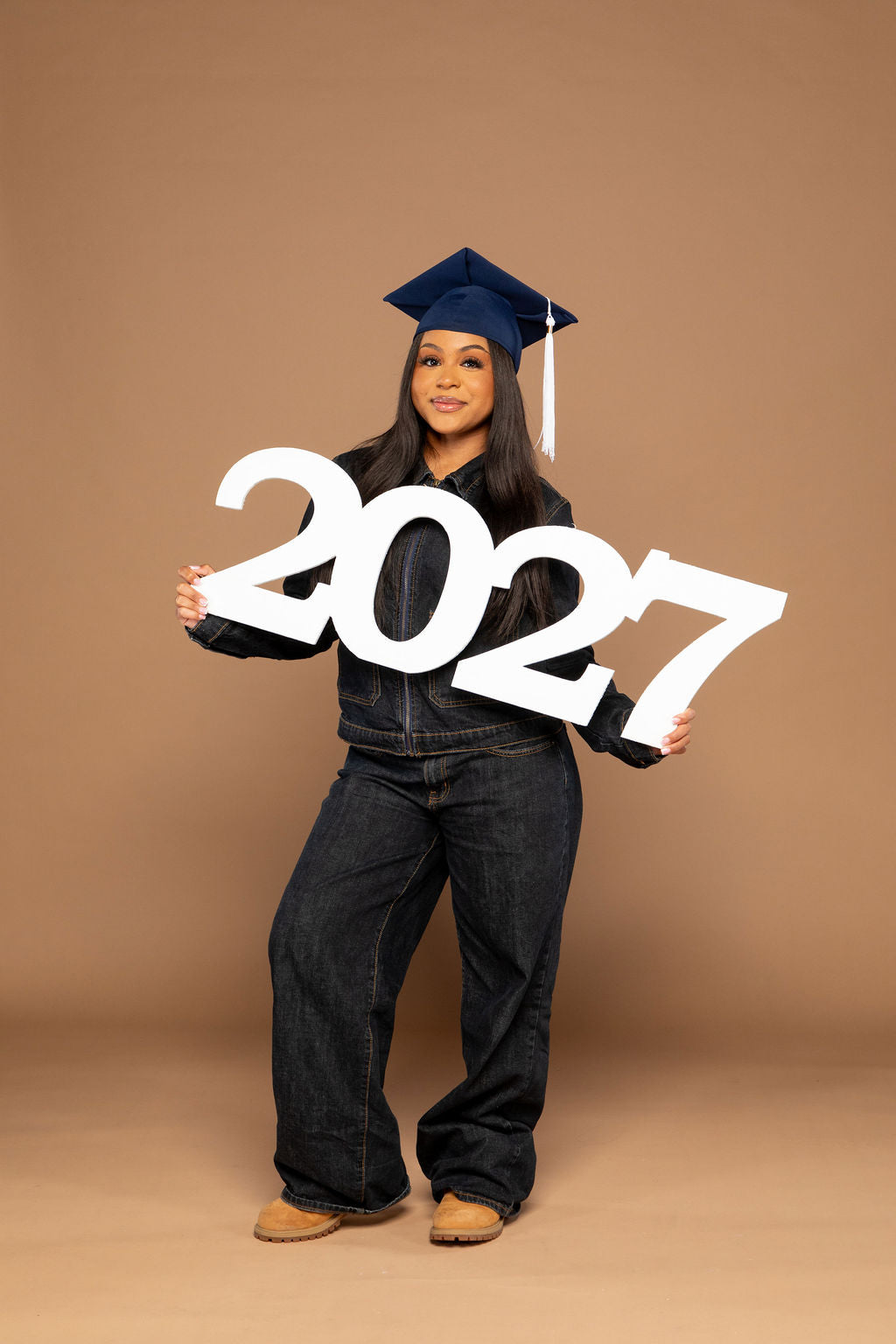 A young woman wearing a navy blue graduation cap and dark jeans stands against a solid tan studio background, holding large white '2027' letter cutouts. Available at collageandwood.com.