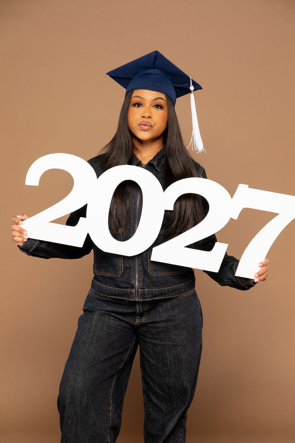 A young woman wearing a navy blue graduation cap and dark denim clothing holds a large white '2027' sign against a solid tan studio background.