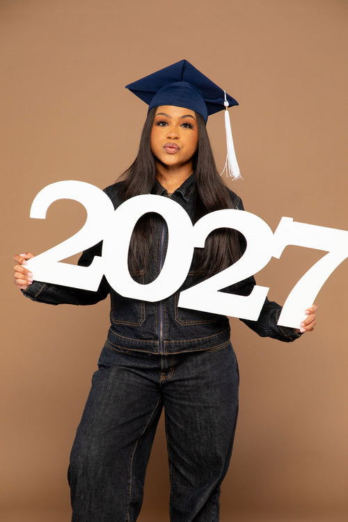 A young woman wearing a navy blue graduation cap and dark denim clothing holds a large white '2027' sign against a solid tan studio background. Available at collageandwood.com.