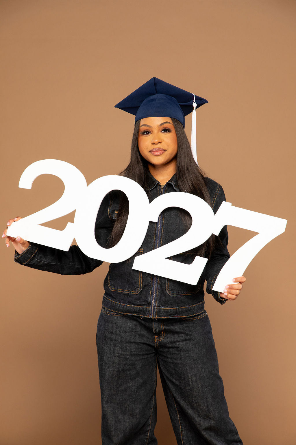 A young woman wearing a navy blue graduation cap and dark denim clothing holds a large white cardboard '2027' sign against a solid brown studio background.