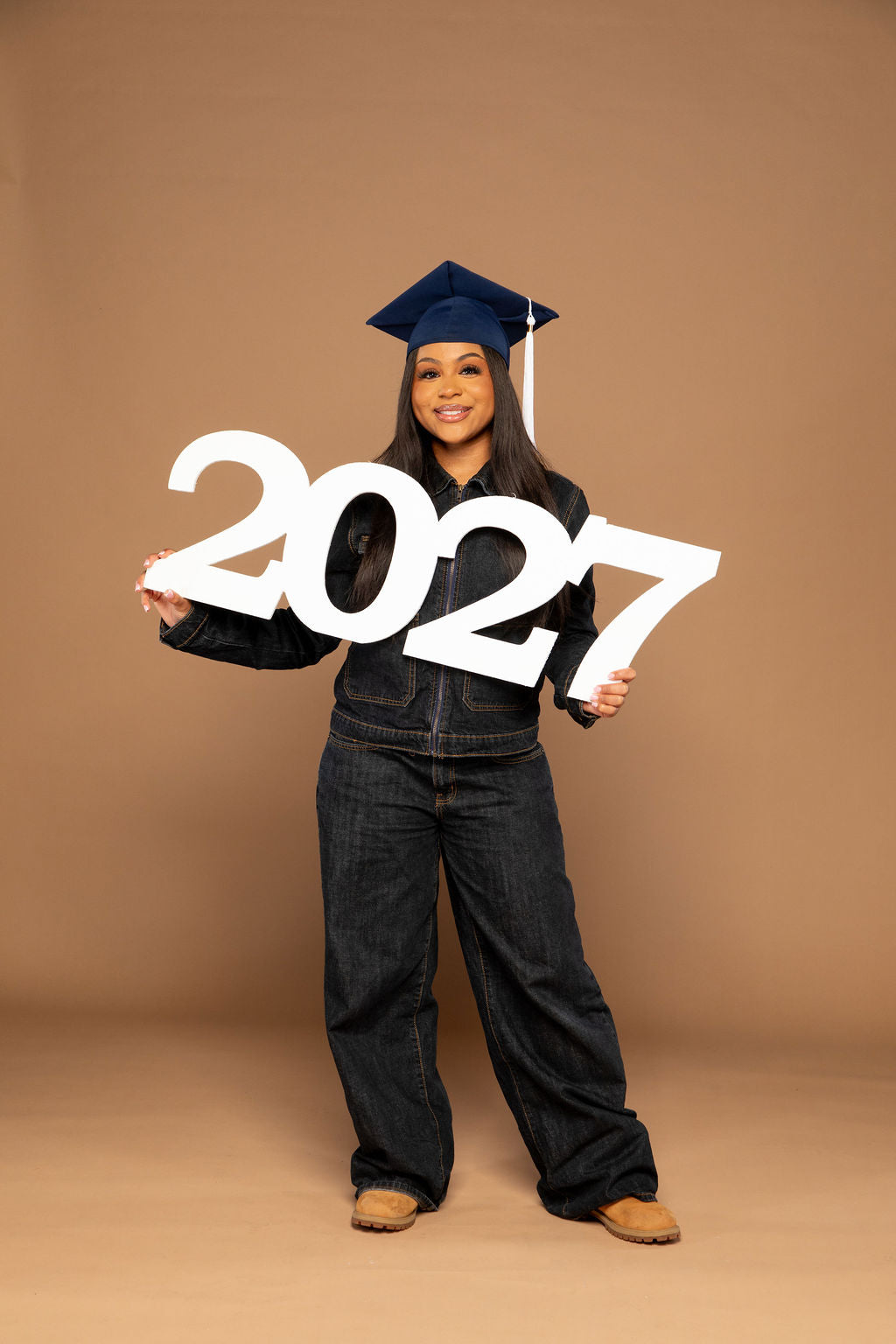 A young woman wearing a navy blue graduation cap and dark denim clothing stands against a solid tan studio backdrop, holding a large white '2027' sign. Available at collageandwood.com.