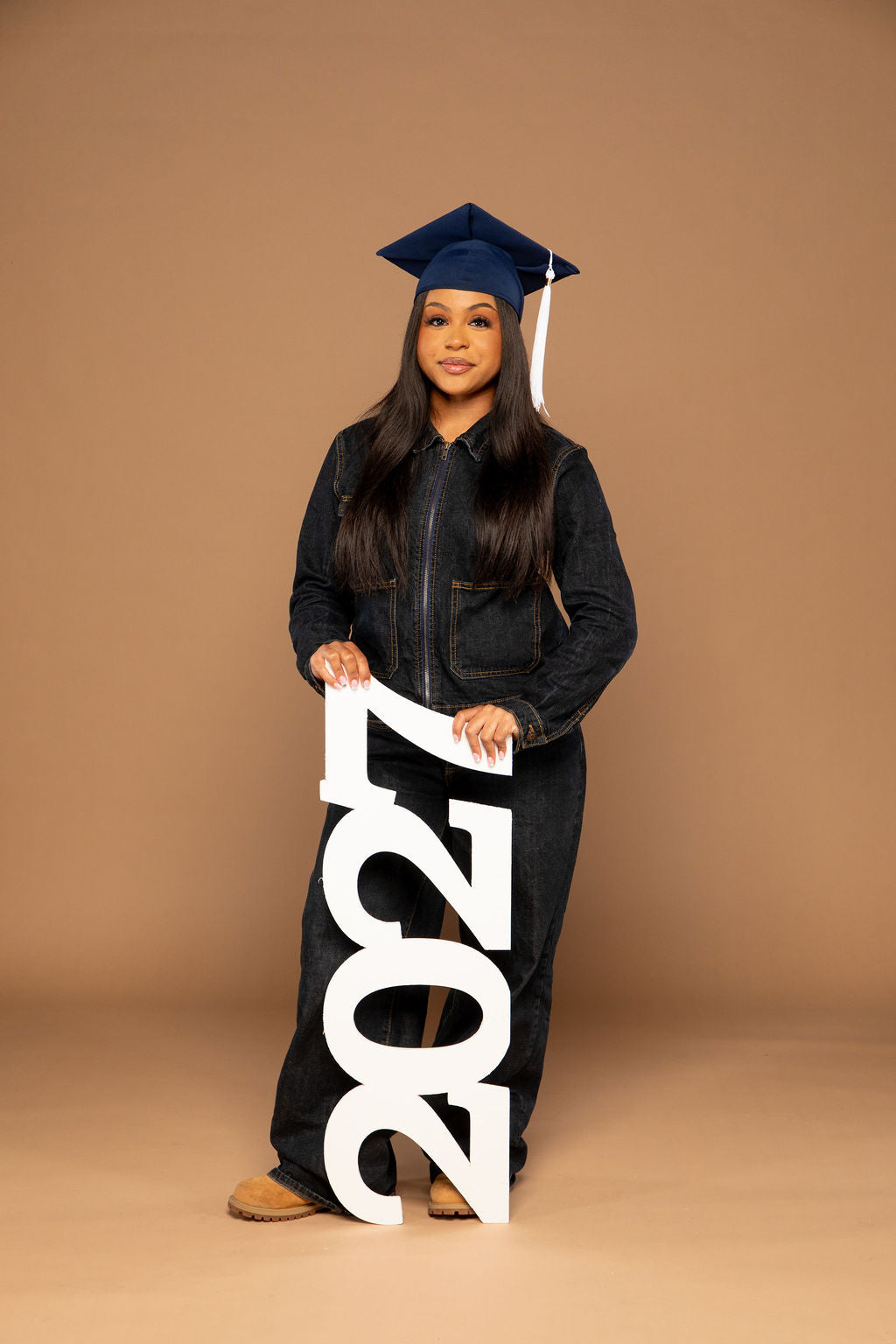 A young woman wearing a navy blue graduation cap and gown stands against a solid tan studio background, holding a large white freestanding '2027' sign.
