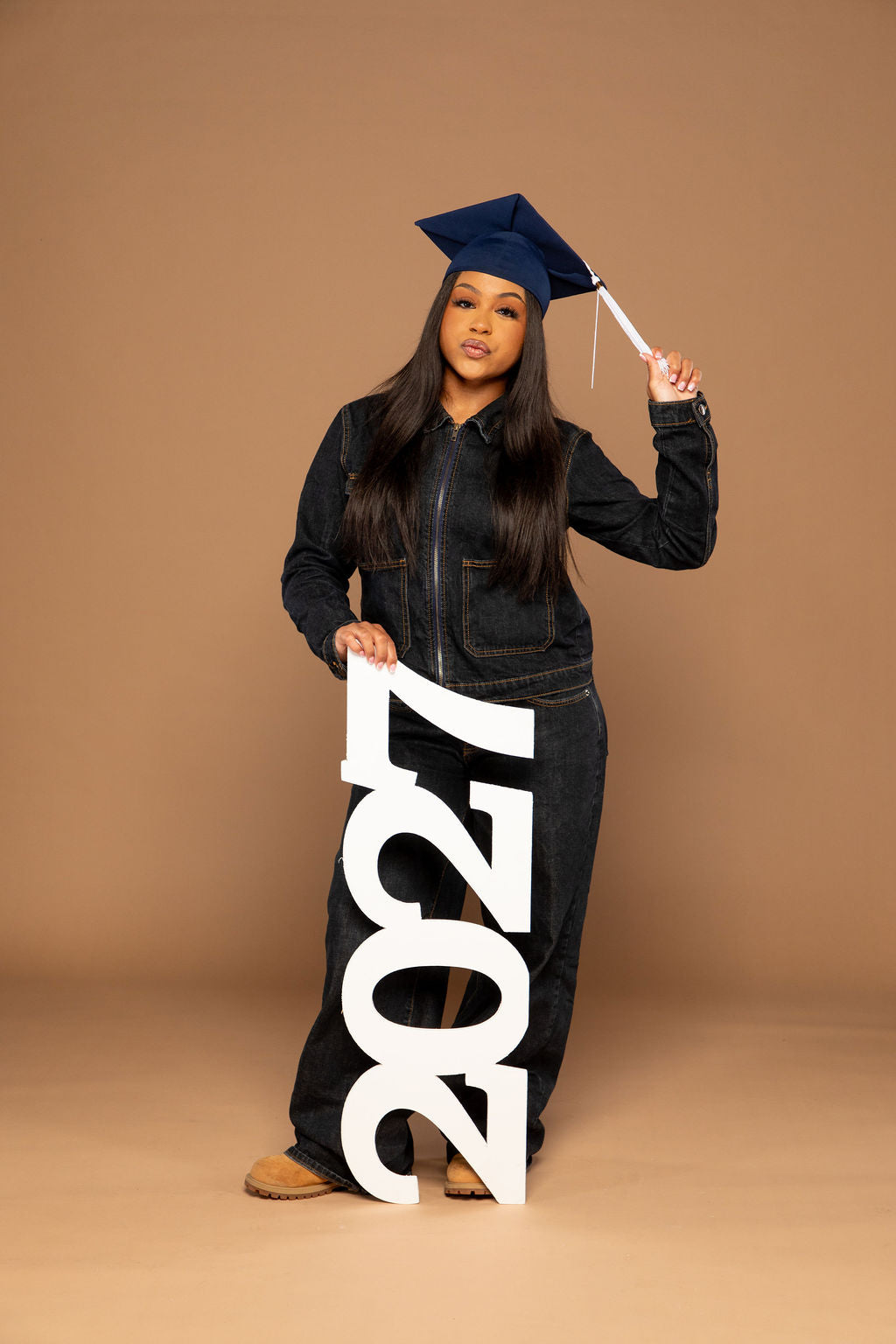 A young woman stands in a studio wearing a dark denim jumpsuit, a navy blue graduation cap, and holding a large white '2027' sign. The background is a solid warm tan color.