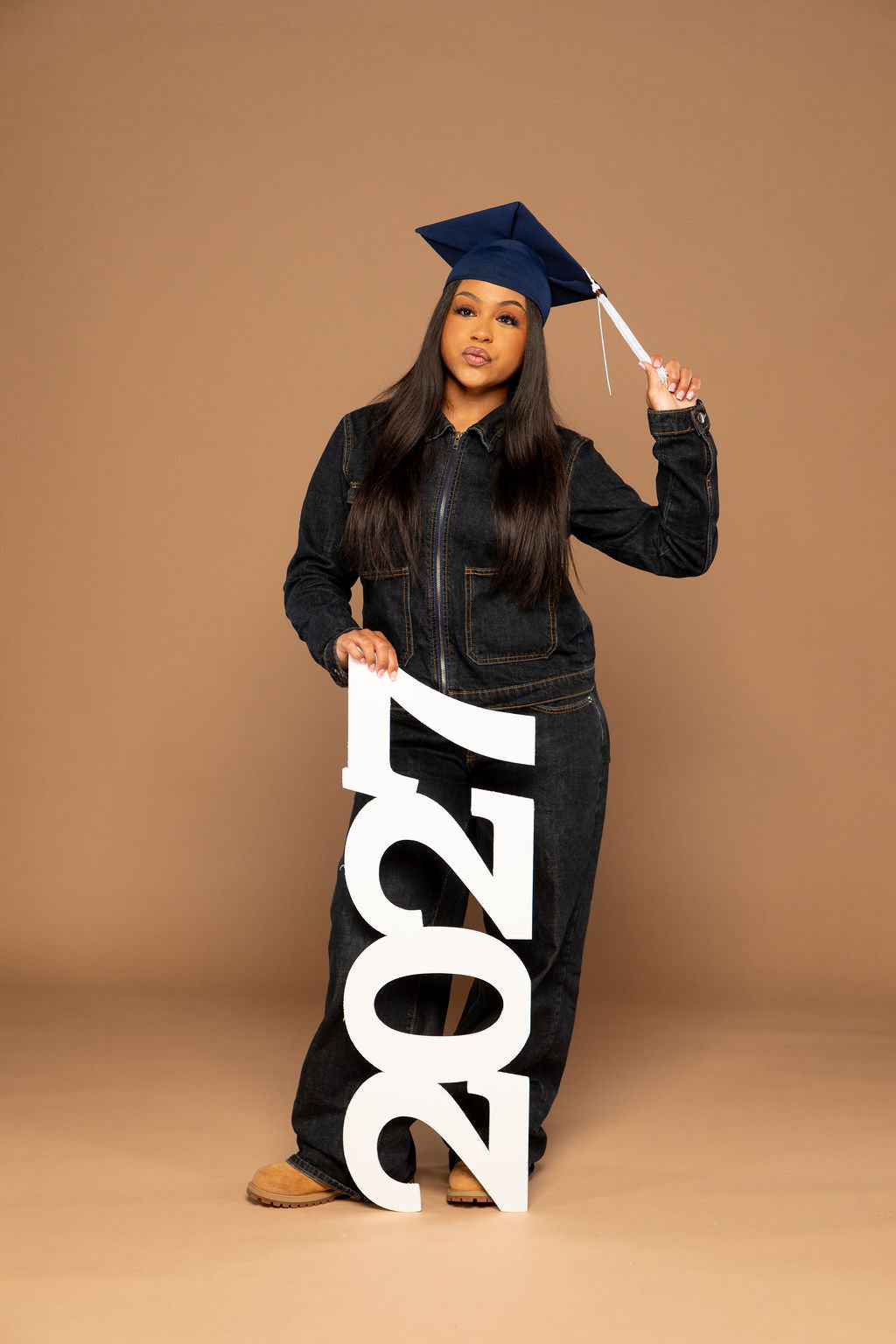 A young woman wearing a dark denim jumpsuit and a navy blue graduation cap stands against a solid tan studio background, holding a large white '2027' sign. She is holding the sign with one hand and touching the brim of her cap with the other.