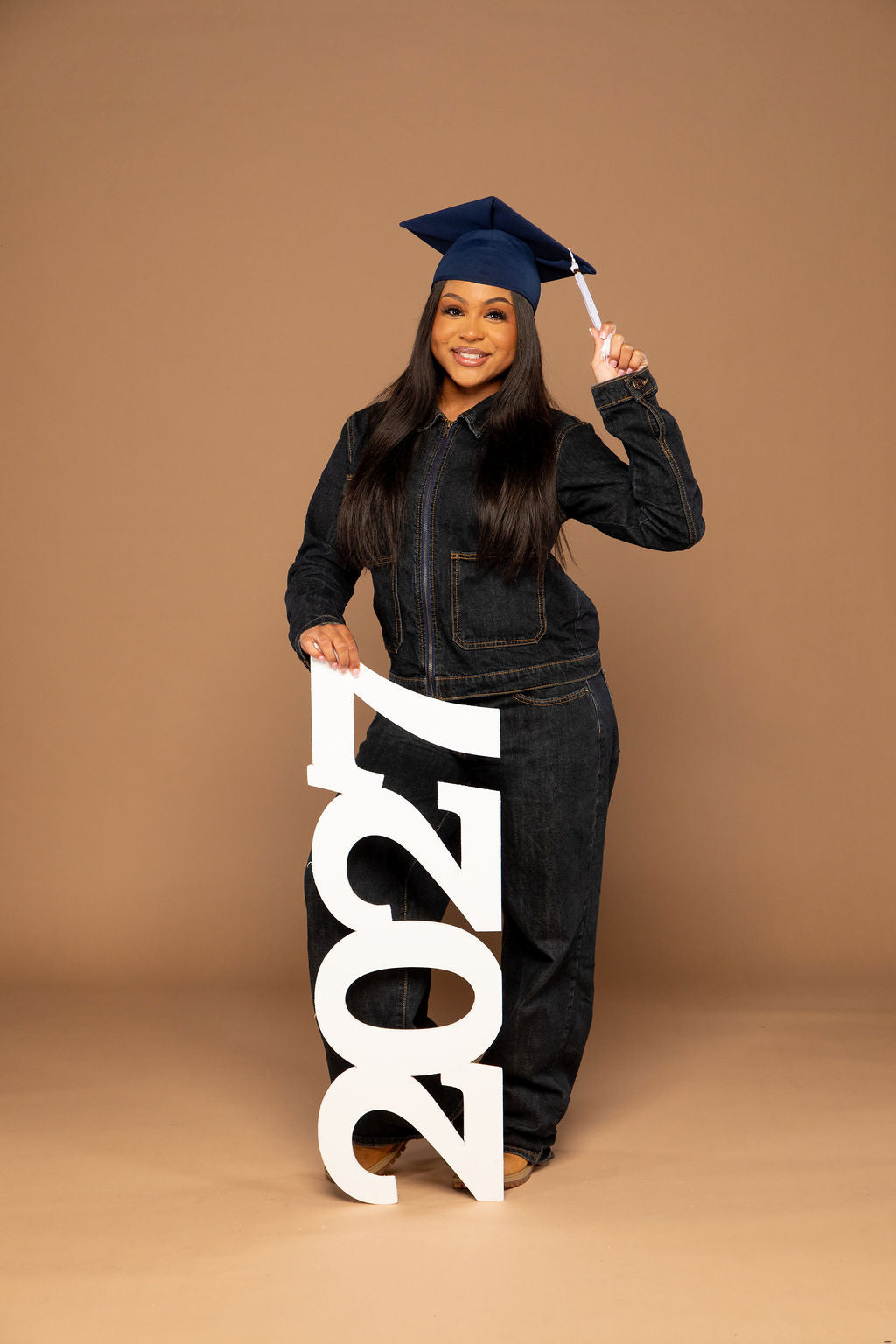 A young woman wearing a dark denim jumpsuit and a navy blue graduation cap stands against a solid tan studio background, holding a large white freestanding '2027' sign. She is smiling and holding the cap with one hand.