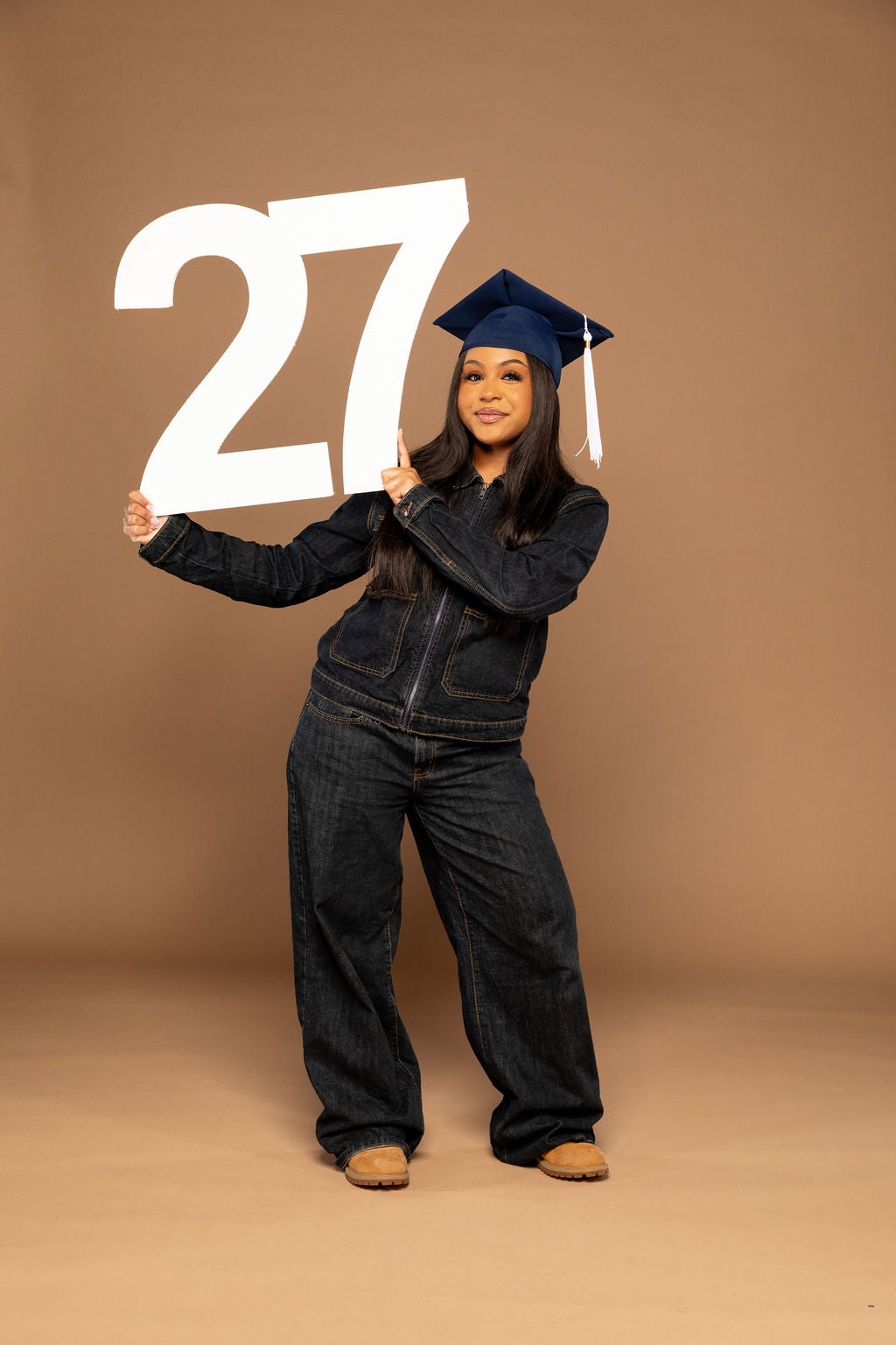 A woman in a dark denim jumpsuit and matching graduation cap holds a large white '27' sign against a solid tan studio backdrop. Available at collageandwood.com.