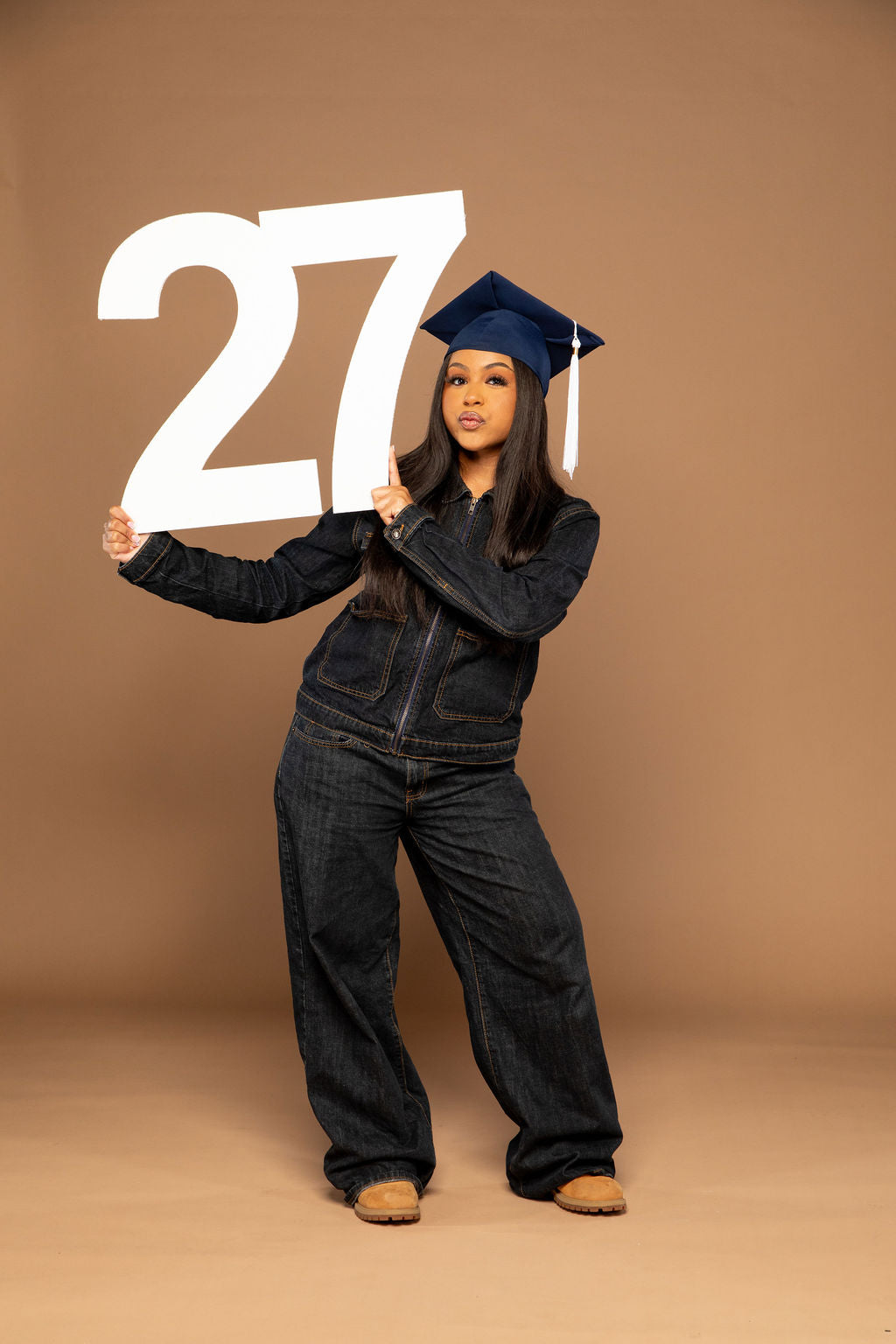 A woman wearing a dark denim jumpsuit and a navy blue graduation cap holds a large white '27' sign against a solid tan studio background. Available at collageandwood.com.