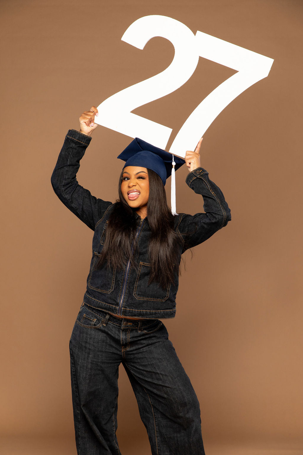 A woman wearing a dark denim jacket and jeans stands against a solid brown studio background, holding a large white '27' sign above her head while wearing a blue graduation cap.