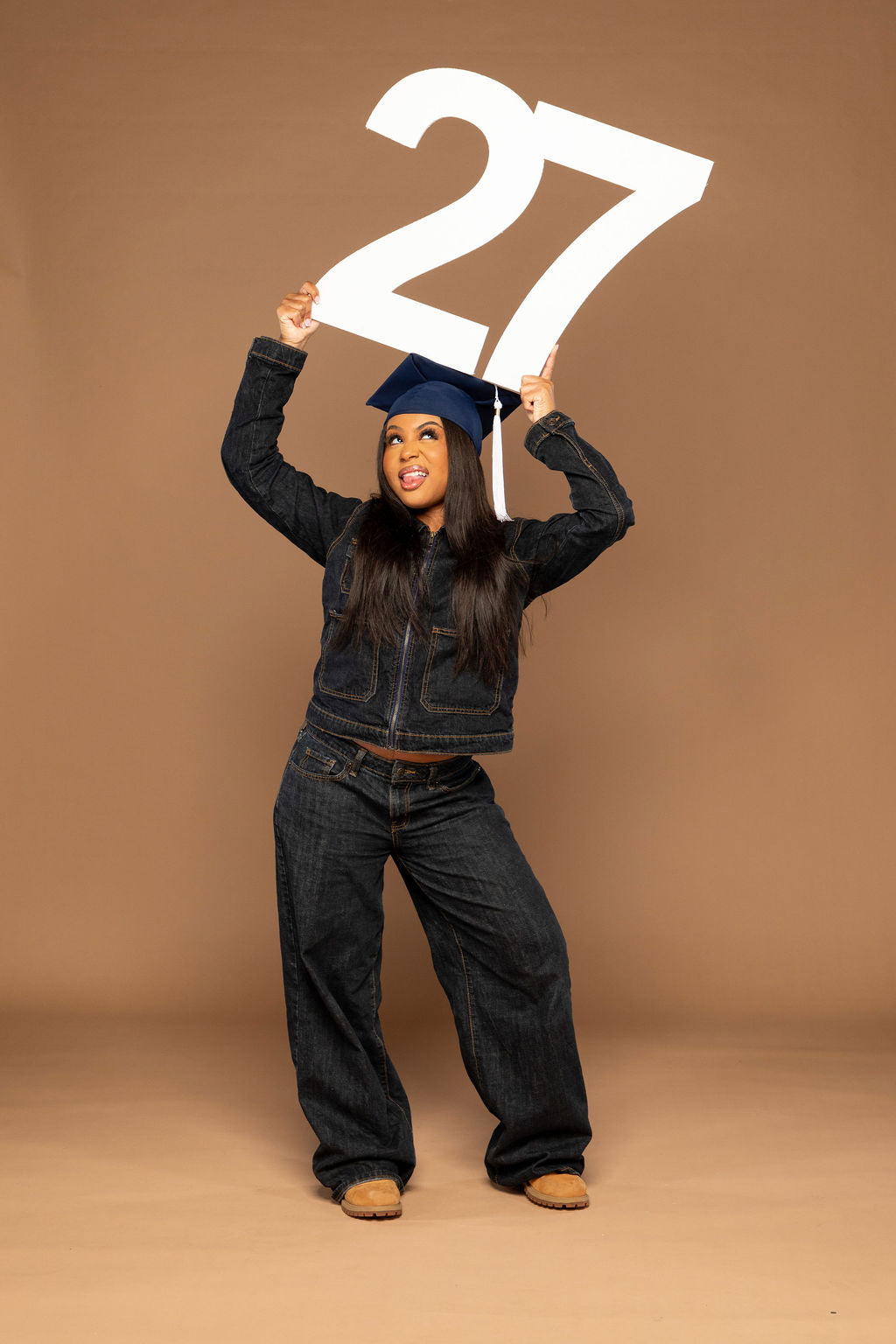 A woman in a dark denim jacket, wide-leg jeans, and a navy blue graduation cap holds a large white '27' sign above her head against a solid tan studio background.
