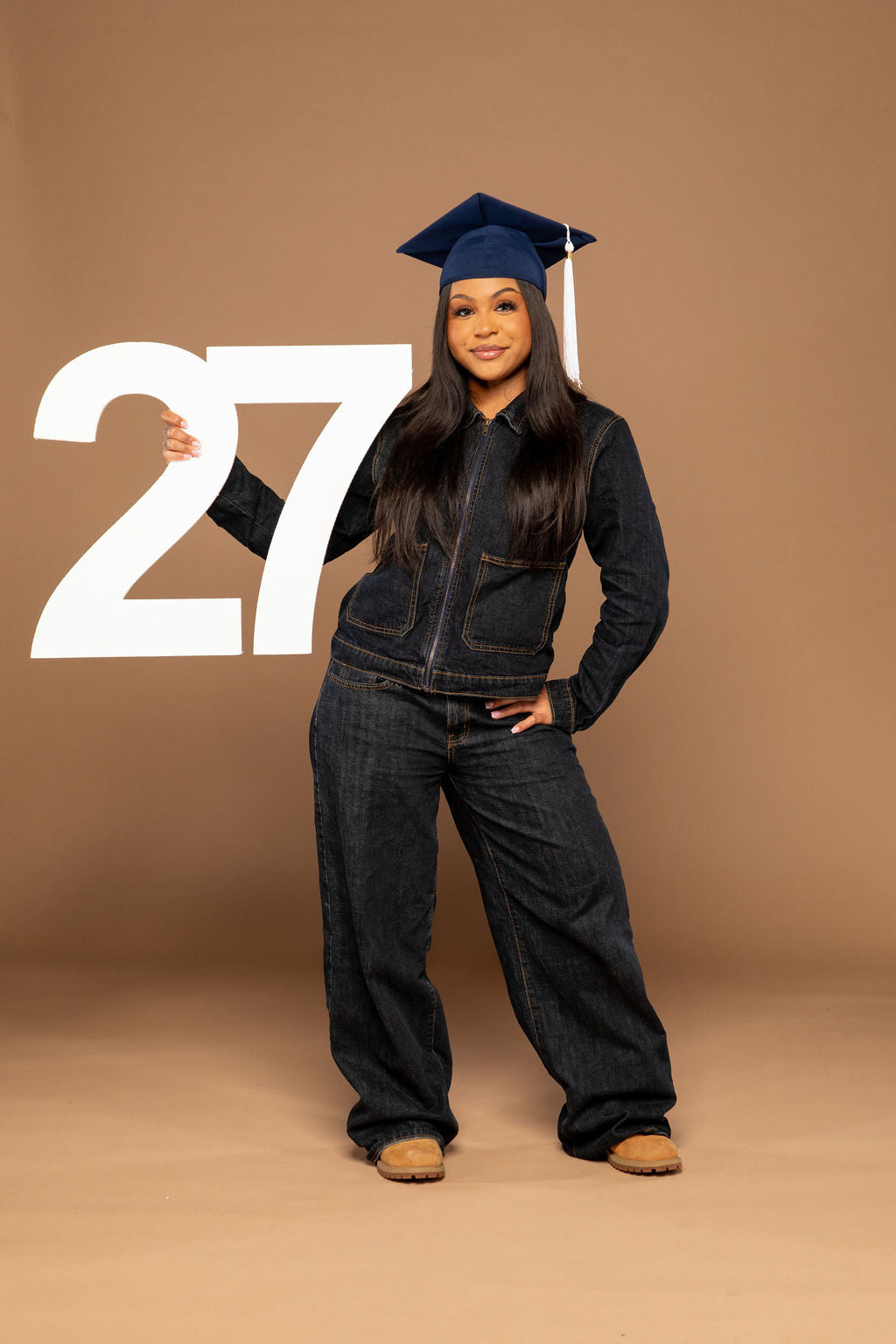 A woman stands against a solid tan studio backdrop wearing a dark denim zip-front jacket and matching wide-leg jeans, with a navy blue graduation cap. She is holding a large white number 27.