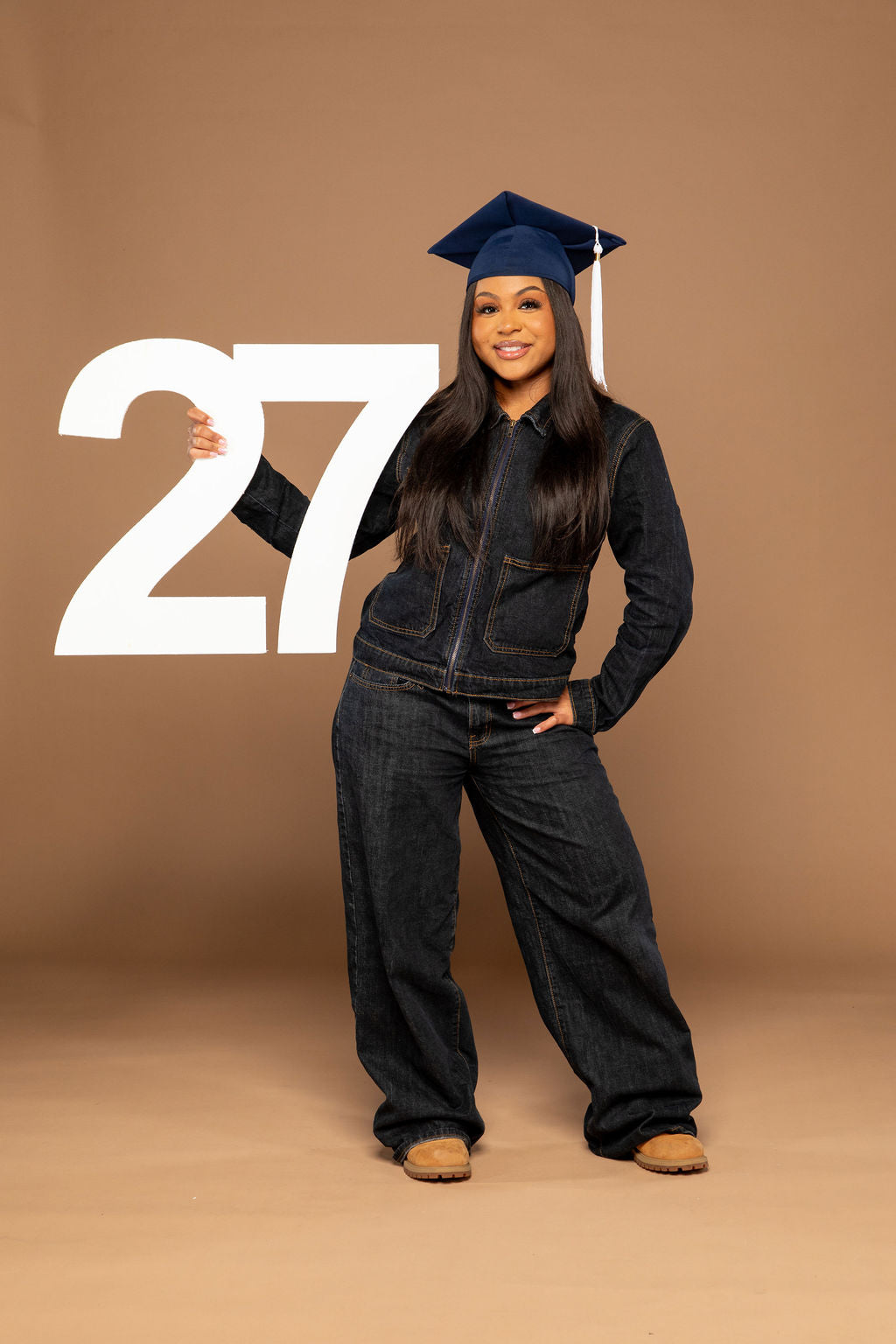 A woman wearing a dark denim jumpsuit, dark blue graduation cap, and tan shoes stands against a solid tan studio background, holding a large white number 27. Available at collageandwood.com.