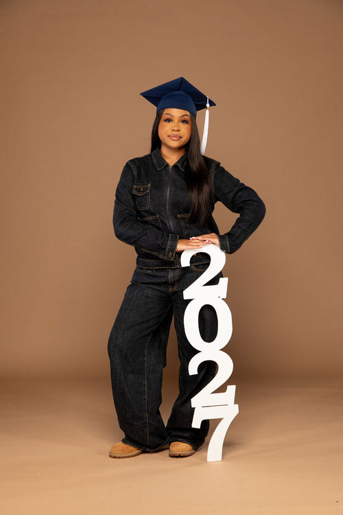 A young woman wearing a dark denim jumpsuit, dark blue graduation cap, and tan shoes stands against a solid brown studio background, holding a large white freestanding '2027' decoration.