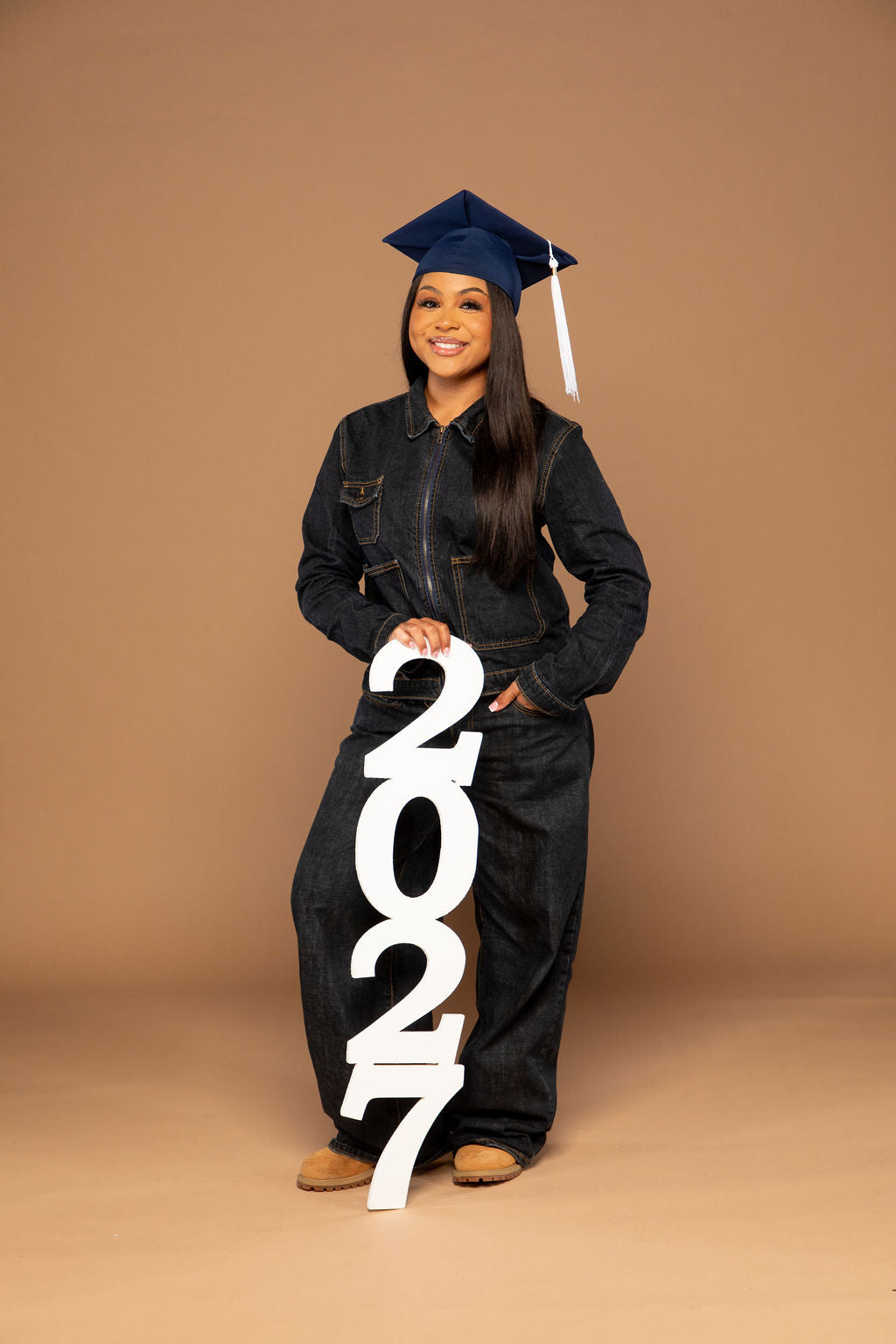 A young woman wearing a dark denim jumpsuit, a navy blue graduation cap, and brown shoes stands against a solid tan studio background, holding a large white '2027' sign.