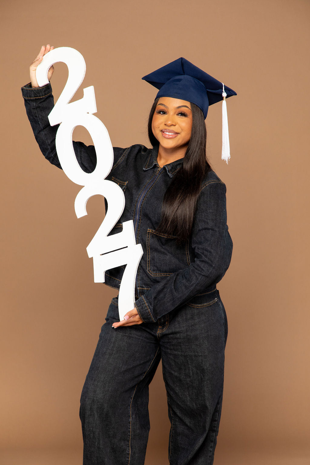 A young woman wearing a navy blue graduation cap and dark denim clothing holds a large white '2024' sign against a solid tan studio background.