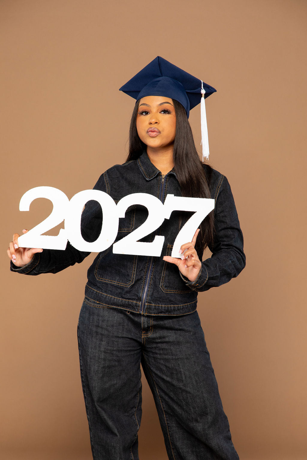 A woman wearing a navy blue graduation cap and dark denim clothing holds a large white '2027' sign against a solid tan studio background.