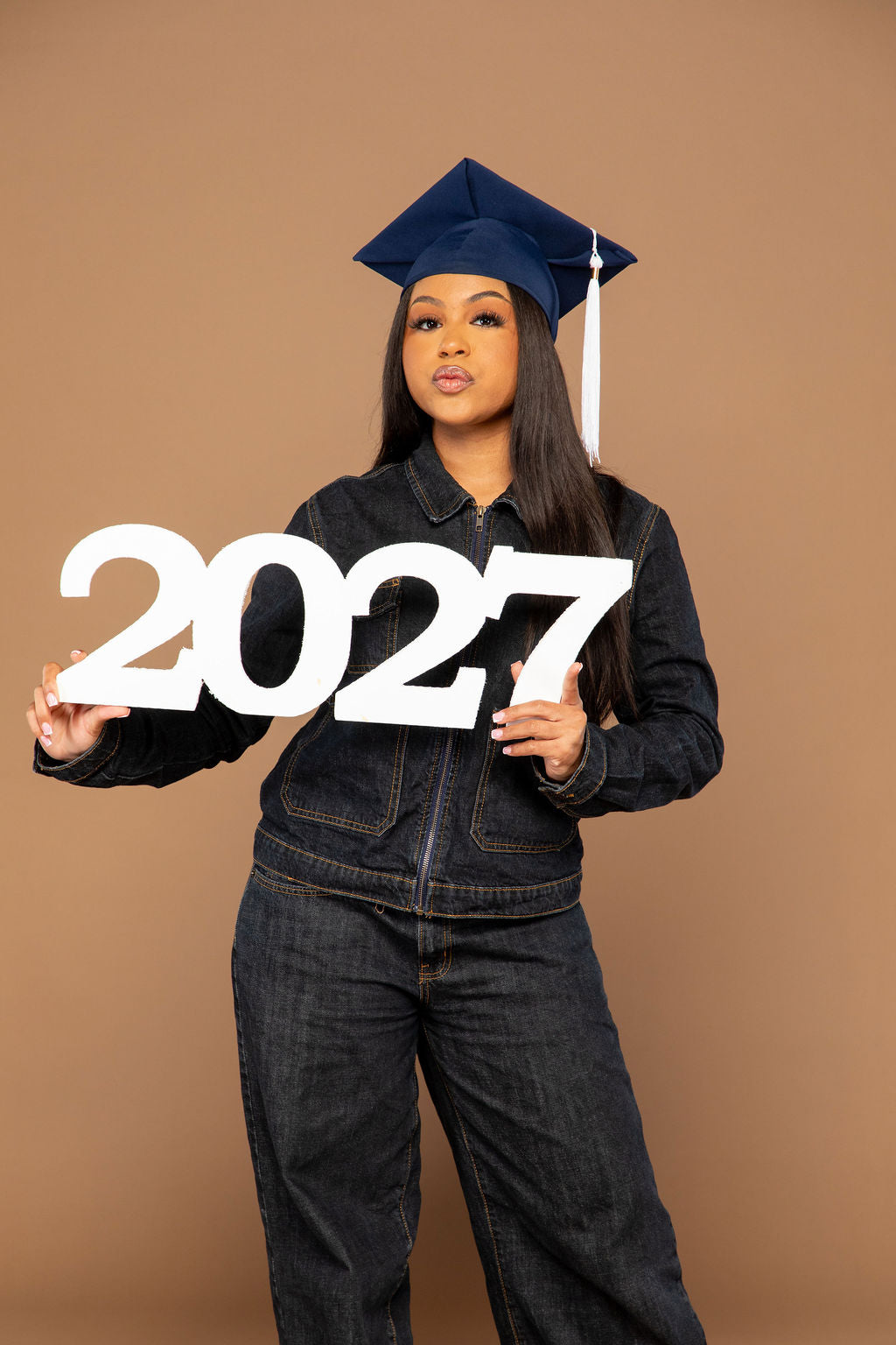 A young woman wearing a navy blue graduation cap and dark denim jacket and jeans stands against a solid tan studio background, holding large white '2027' lettering.