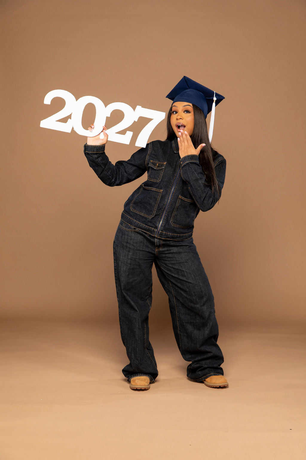 A woman posing in a dark denim jumpsuit and a navy blue graduation cap, holding a white '2027' sign against a solid tan studio backdrop.