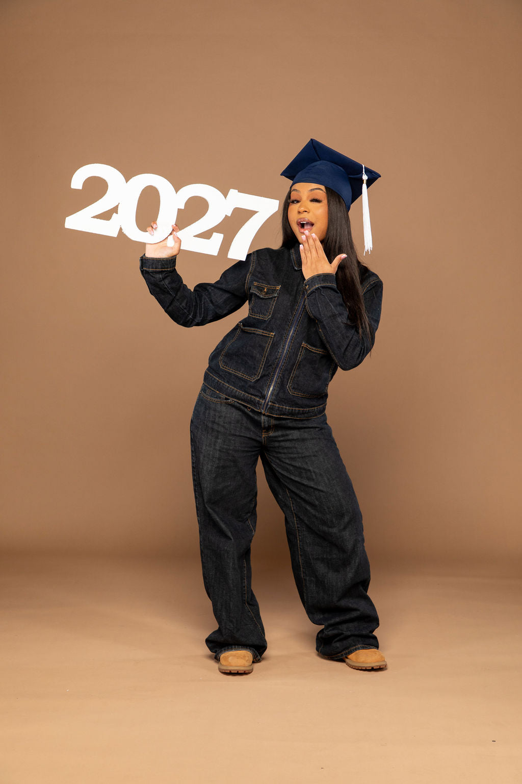 A young woman wearing a dark denim jumpsuit and a navy blue graduation cap poses with a large white '2027' sign against a solid tan studio backdrop.