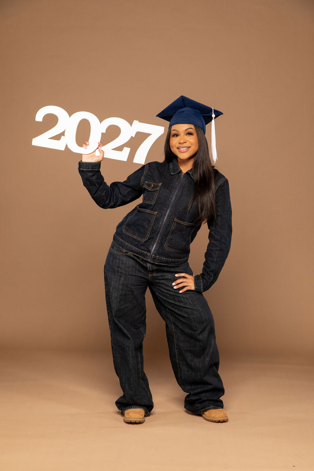 A young woman wearing a dark denim jumpsuit and a navy blue graduation cap poses with large white '2027' numbers against a solid tan studio backdrop.