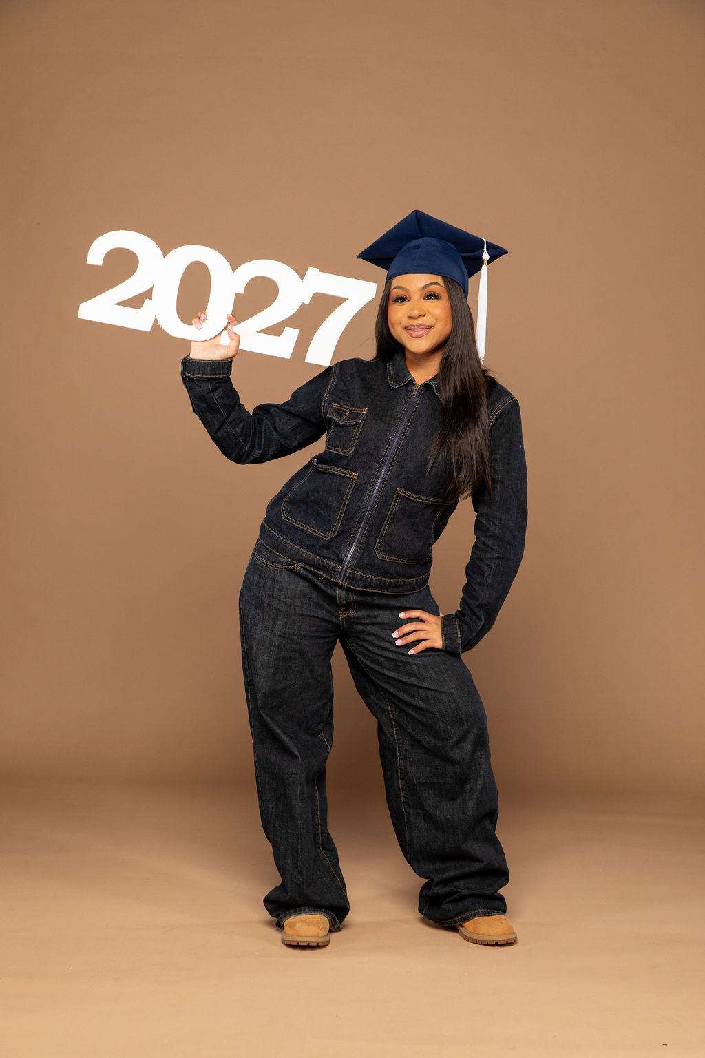 A young woman wearing a dark denim jumpsuit, matching dark jeans, tan boots, and a navy blue graduation cap stands against a solid tan studio background, holding up large white '2027' letters.