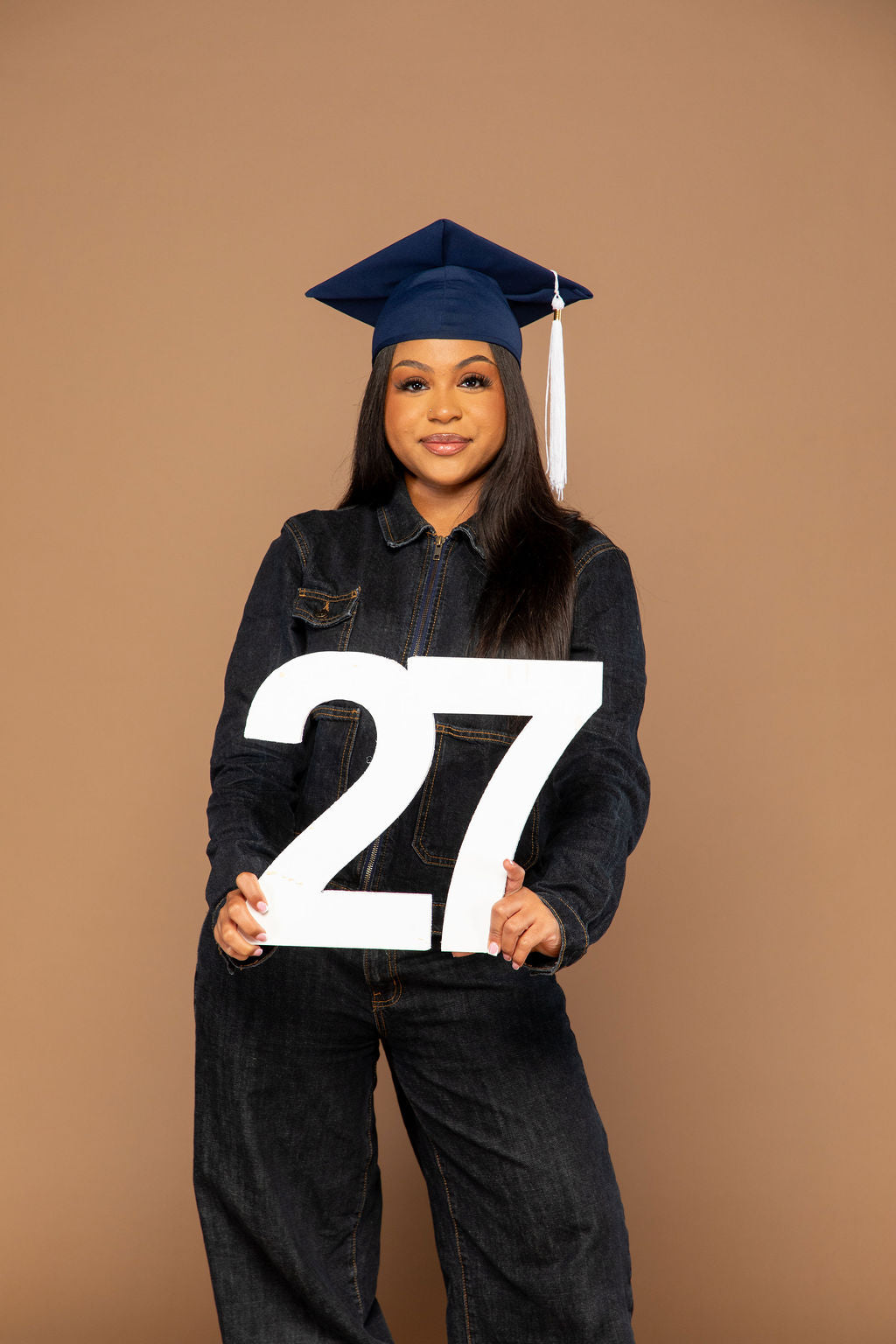 A woman wearing a navy blue graduation cap and dark denim overalls holds a large white '27' sign against a solid tan studio background. Available at collageandwood.com.