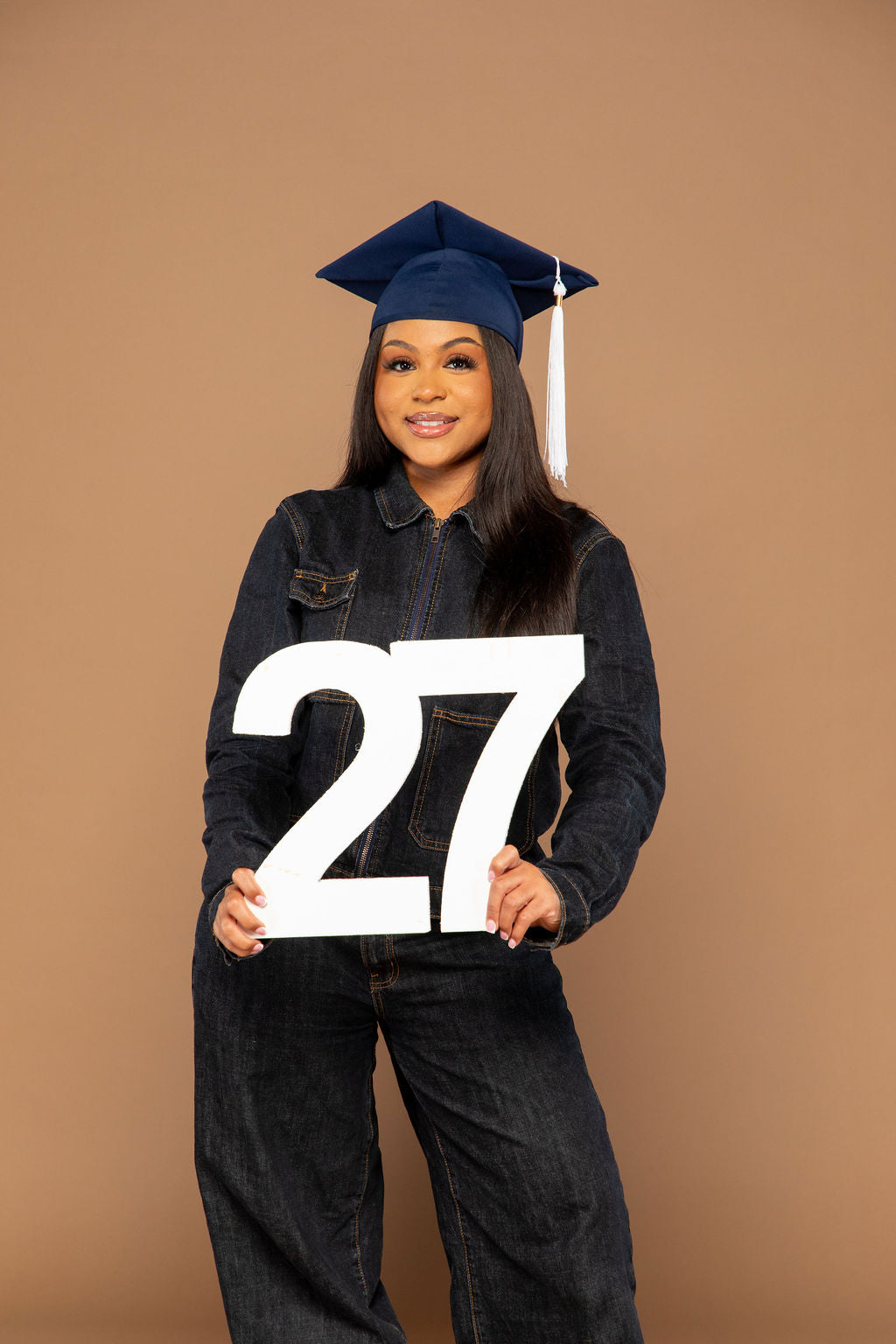A woman wearing a navy blue graduation cap and matching denim outfit stands against a solid tan background, holding a large white '27' sign. Available at collageandwood.com.