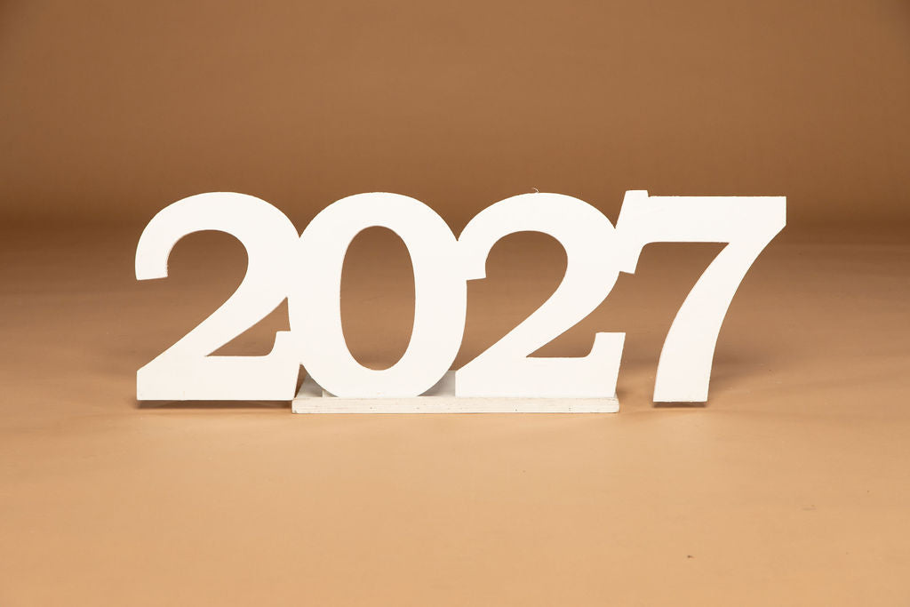 White 3D cutout numbers '2027' standing on a small white base against a warm brown background.