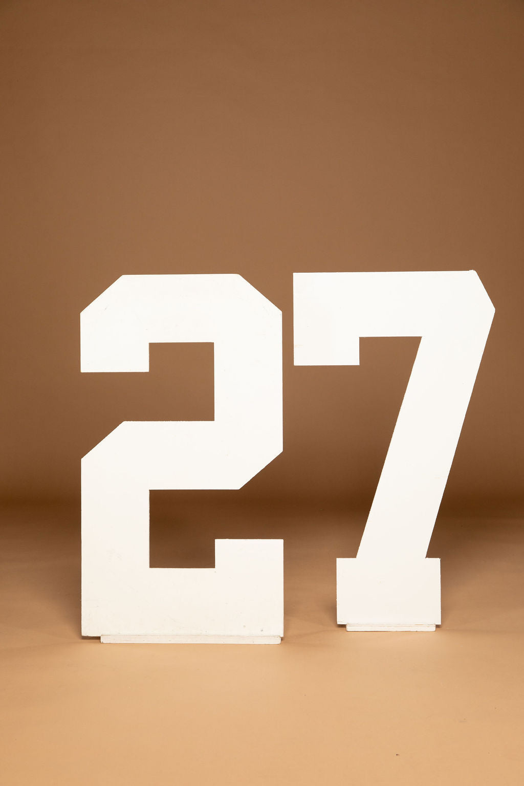 Large white freestanding numbers '27' on a warm brown studio background.