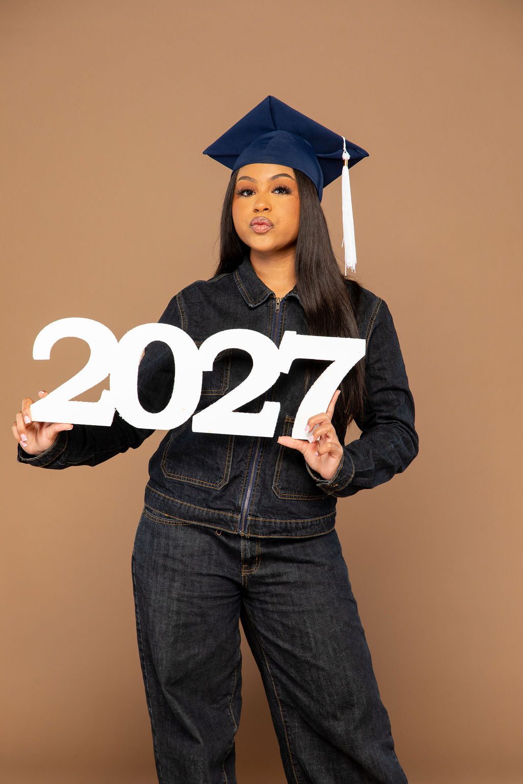 Graduate in a cap holding a white wooden 2027 photo prop