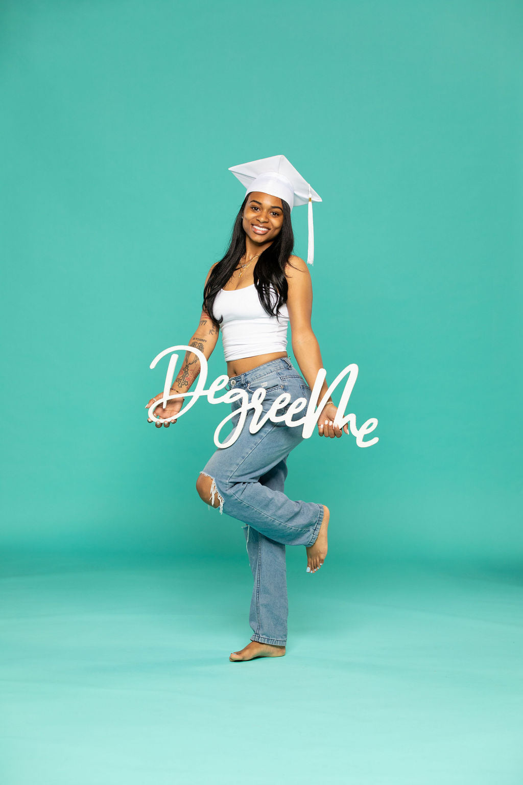 Graduate wearing a white cap stands on teal floor holding a “DegreeMe” prop sign in front of her. Degree Me props available now at collageandwood.com.