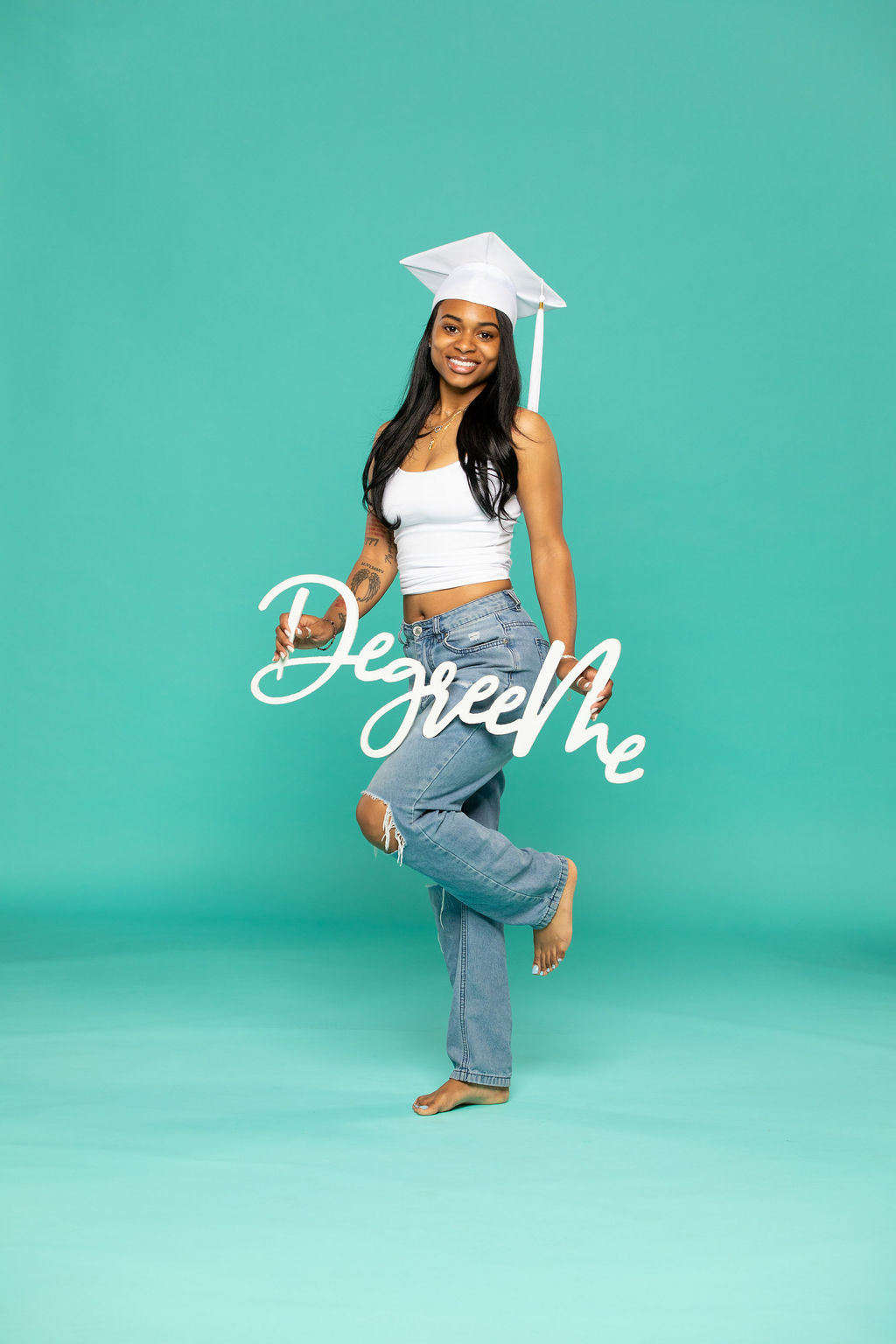 Joyful graduate in white cap and ripped jeans poses on one foot while holding a script sign that says “DegreeMe.” Degree Me props available now at collageandwood.com.