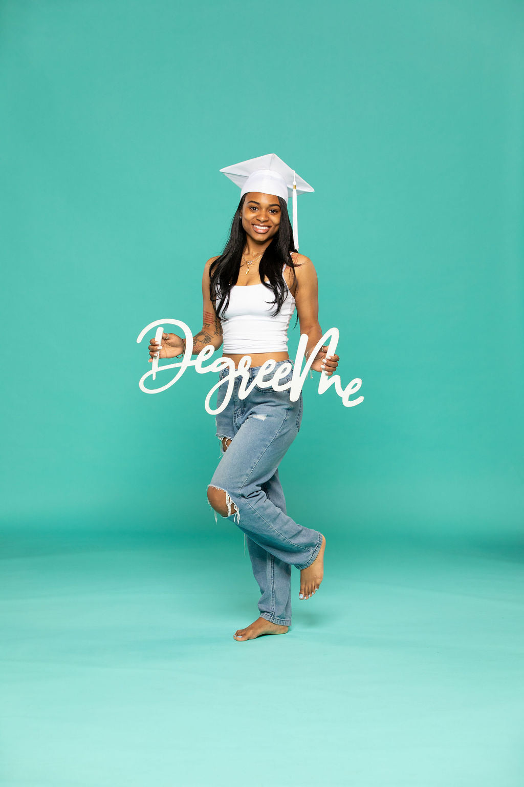 Joyful graduate in white cap and ripped jeans poses on one foot while holding a script sign that says “DegreeMe.” Degree Me props available now at collageandwood.com.