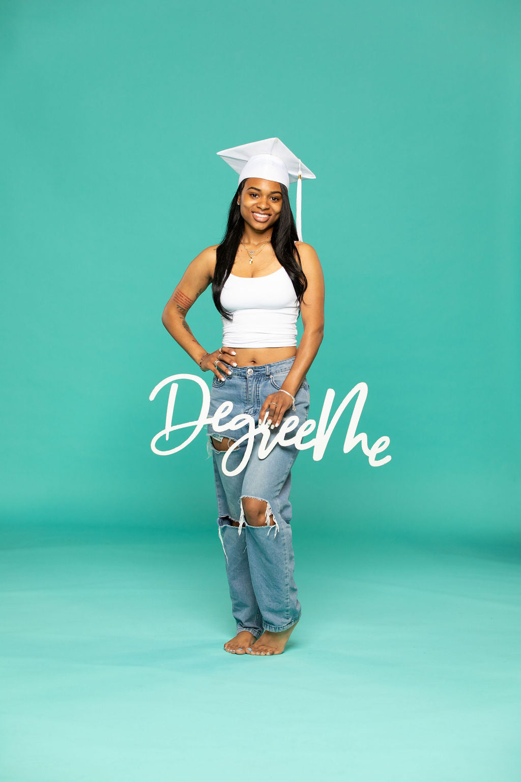 Joyful graduate in white cap and ripped jeans poses on one foot while holding a script sign that says “DegreeMe.” Degree Me props available now at collageandwood.com.
