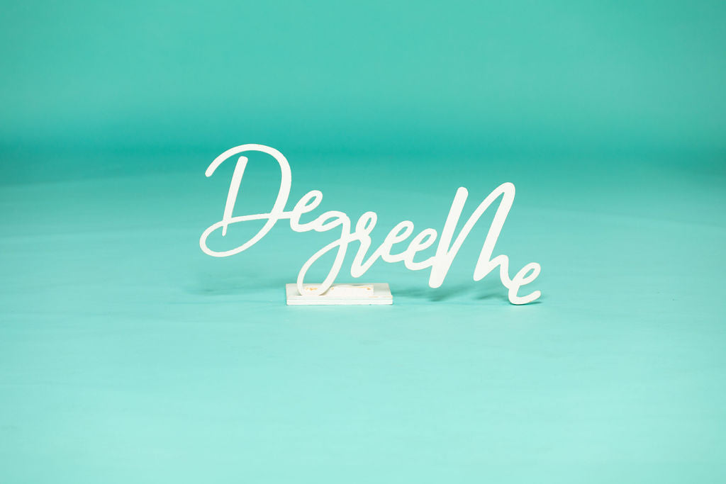 Close-up of a freestanding white “DegreeMe” sign on a teal background, designed as a graduation photo prop. Degree Me props available now at collageandwood.com.