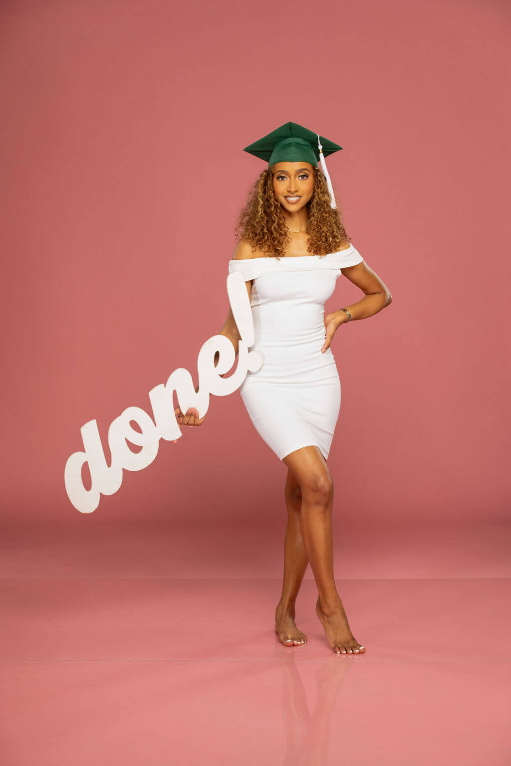 Senior model posing with "done!" photo prop. Model is wearing a white dress in a professional studio with a pink background.