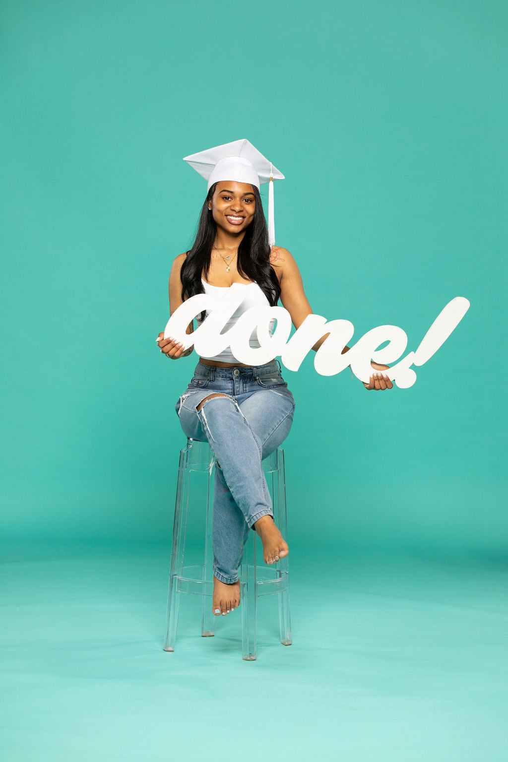 miling graduate in white cap and ripped jeans sits barefoot on a clear stool, proudly holding a script sign that reads “done!” against a teal background. 2026 senior props available now at collageandwood.com.