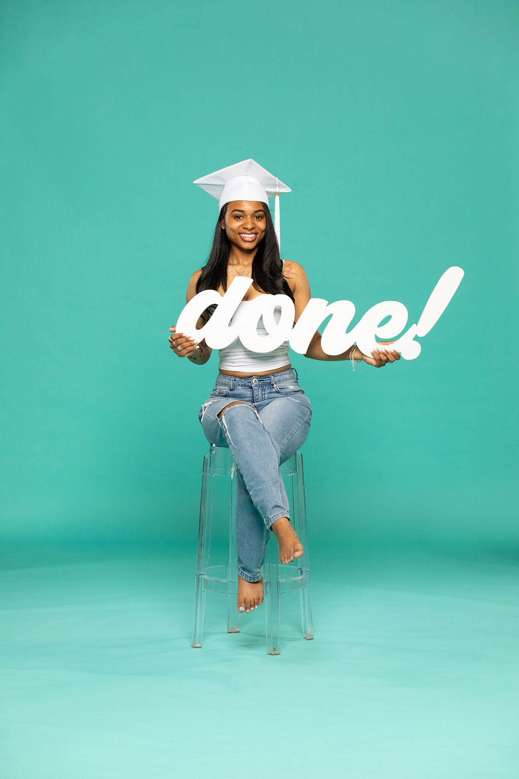 miling graduate in white cap and ripped jeans sits barefoot on a clear stool, proudly holding a script sign that reads “done!” against a teal background. 2026 senior props available now at collageandwood.com.