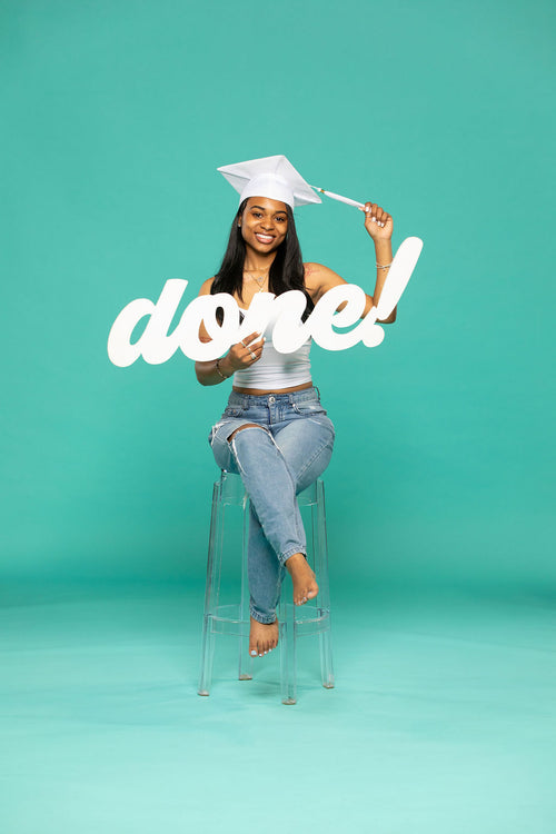 miling graduate in white cap and ripped jeans sits barefoot on a clear stool, proudly holding a script sign that reads “done!” against a teal background. 2026 senior props available now at collageandwood.com.