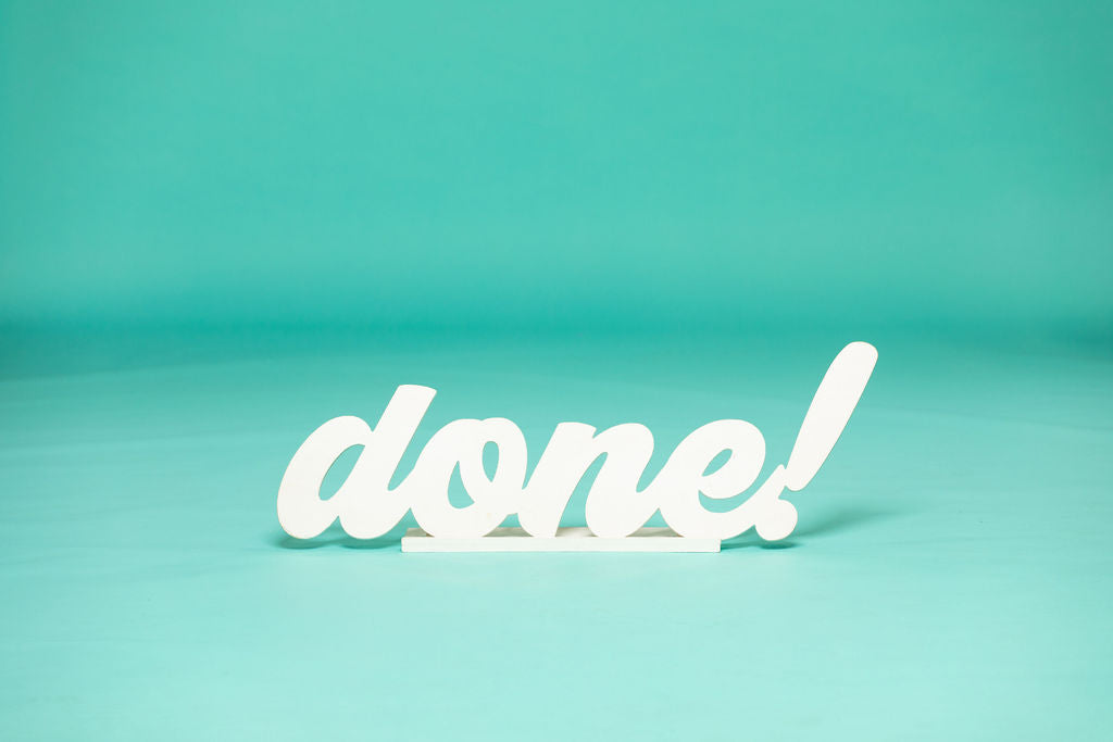 White “done!” sign in bold cursive lettering stands on a teal studio floor, ideal for celebrating graduation milestones. 2026 senior props available now at collageandwood.com.