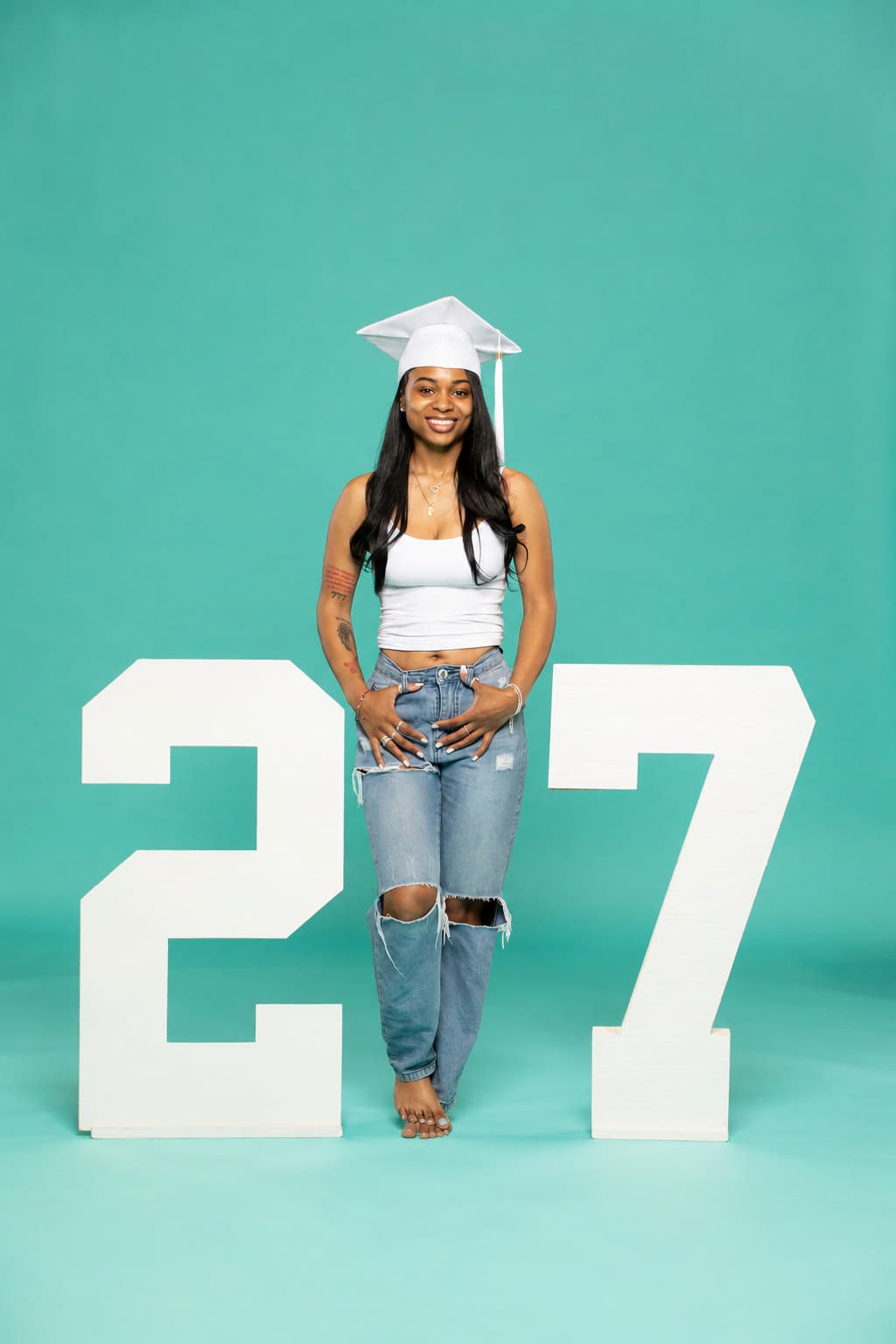 Person in graduation cap and gown standing between large numbers '27' on a teal background. Available at collageandwood.com.