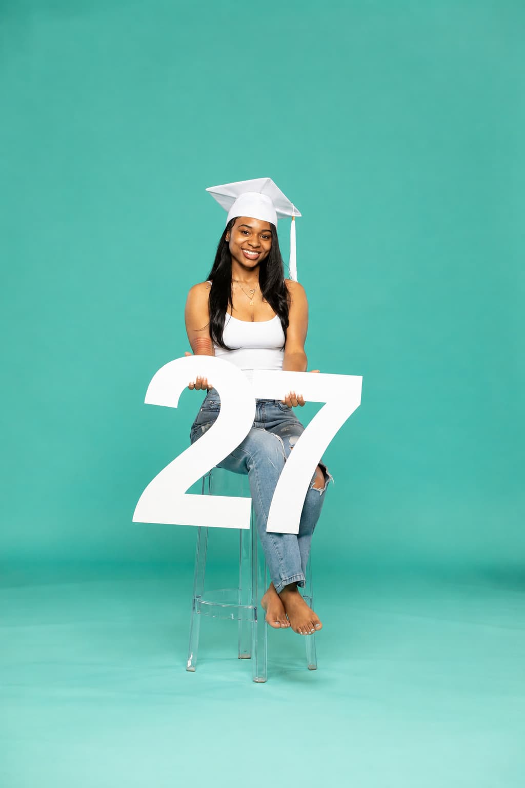 Young woman in a graduation cap sitting barefoot on a clear stool, holding a large white number "27" against a teal background. 2027 senior props available now at collageandwood.com.